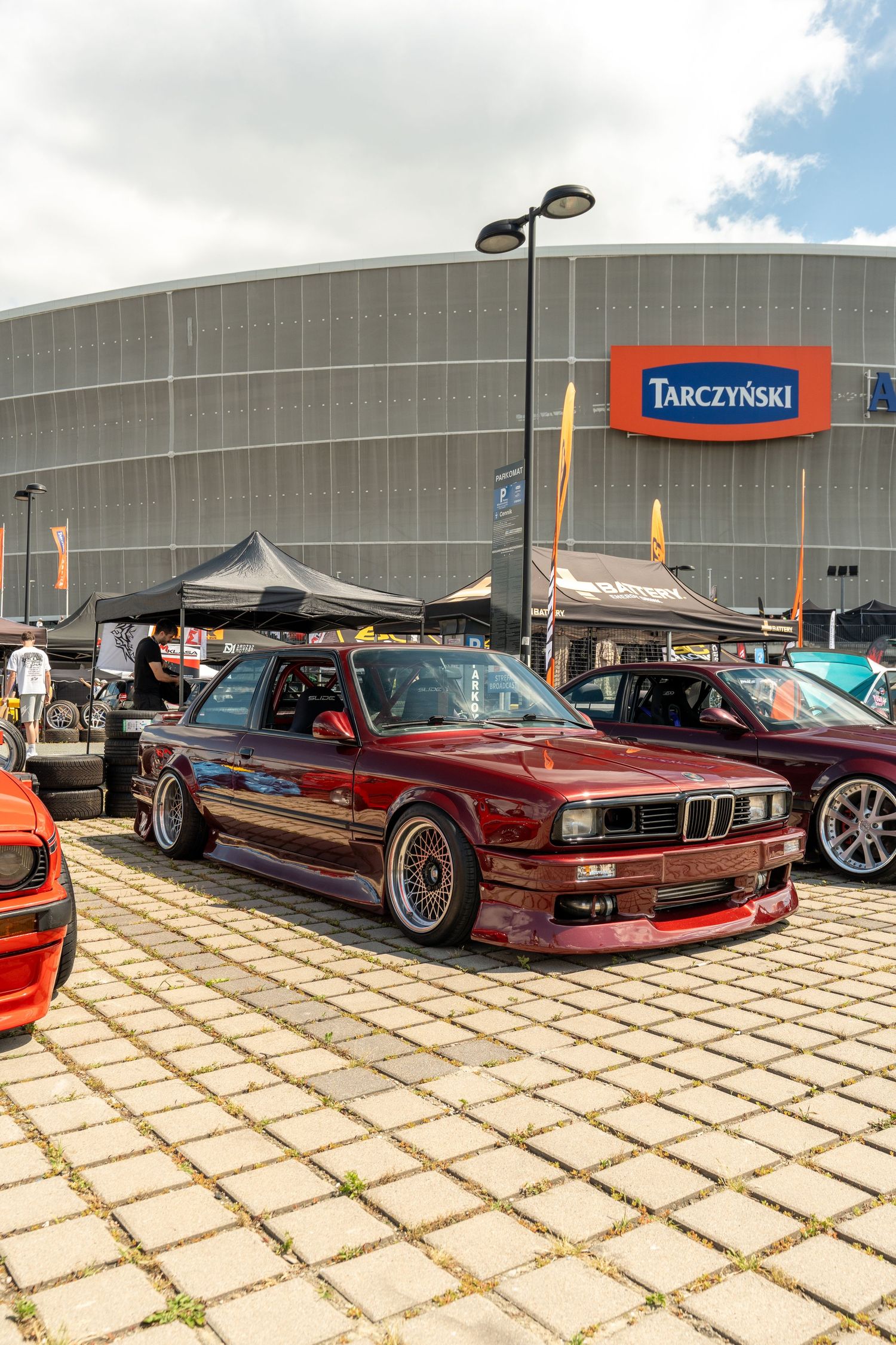 A burgundy modified BMW M3 E30 sits on bronze wheels at a car show in front of Tarczynski building.