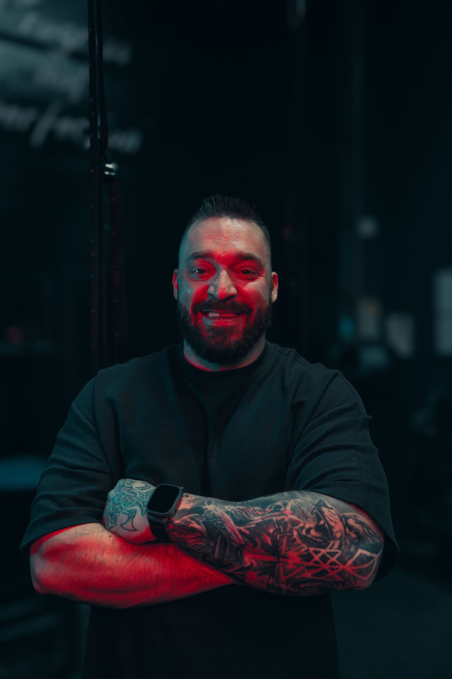 Tattooed person in black sweater smiling with red lighting effect in dark setting.