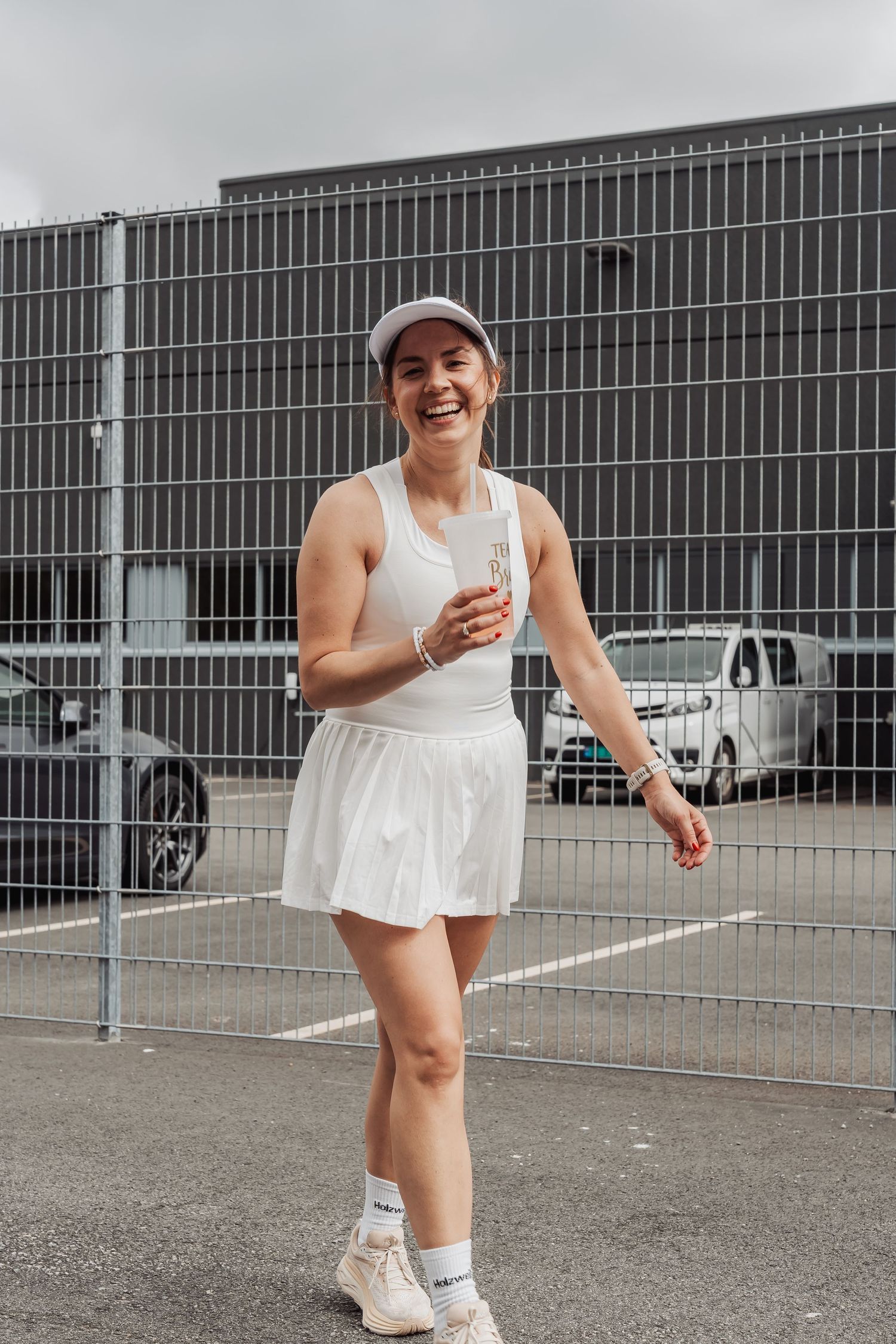 Tennis player in white athletic dress smiling on outdoor court next to metal fence.