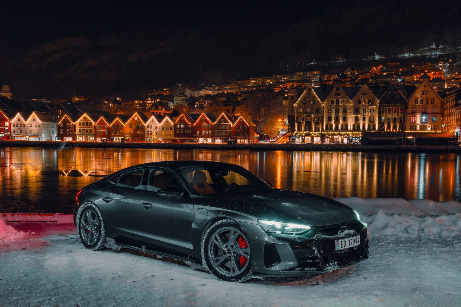 Audi RS e-tron GT sports car parked at night against illuminated waterfront buildings with reflections on water.