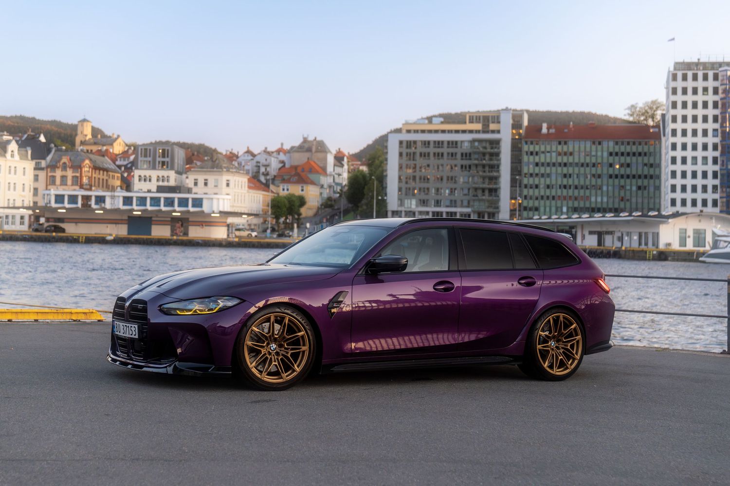A purple BMW M3 wagon with gold wheels parked by a harbor during golden hour.