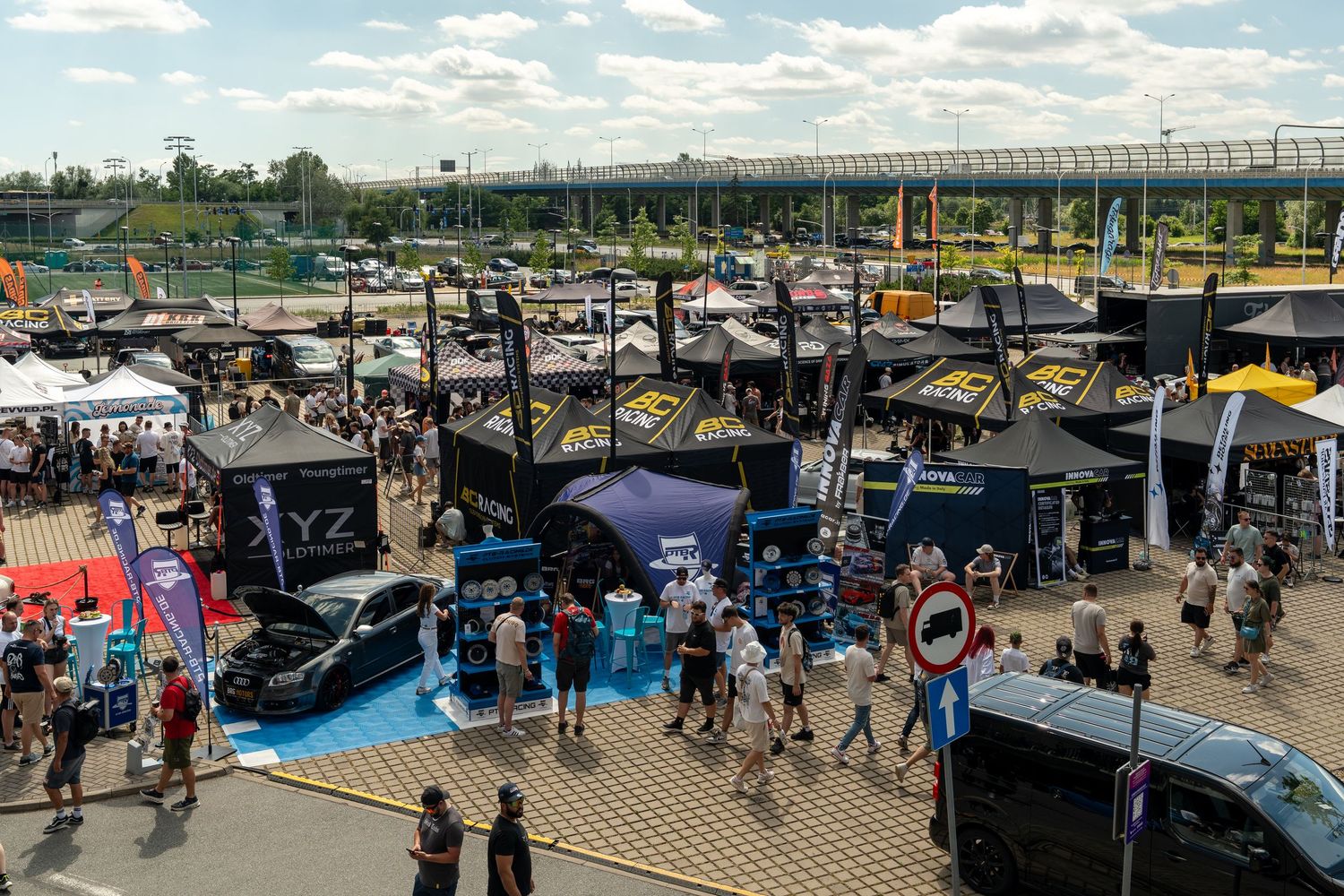 Aerial view of an outdoor car show with various vehicles and vendor tents under blue skies.