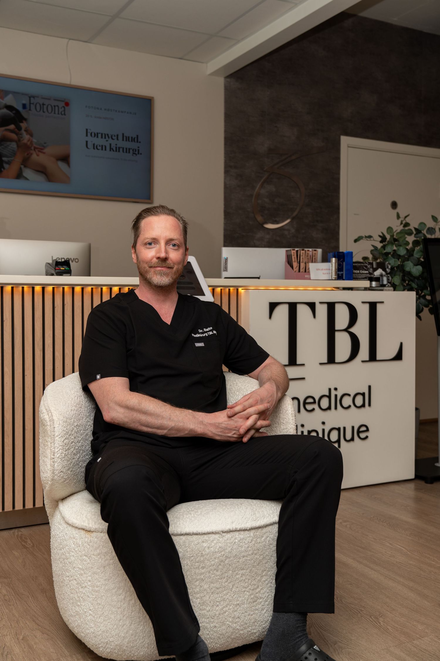 Man in black scrubs sits in a white boucle armchair at the TBL Médical Clinique reception desk in a modern clinic lobby.