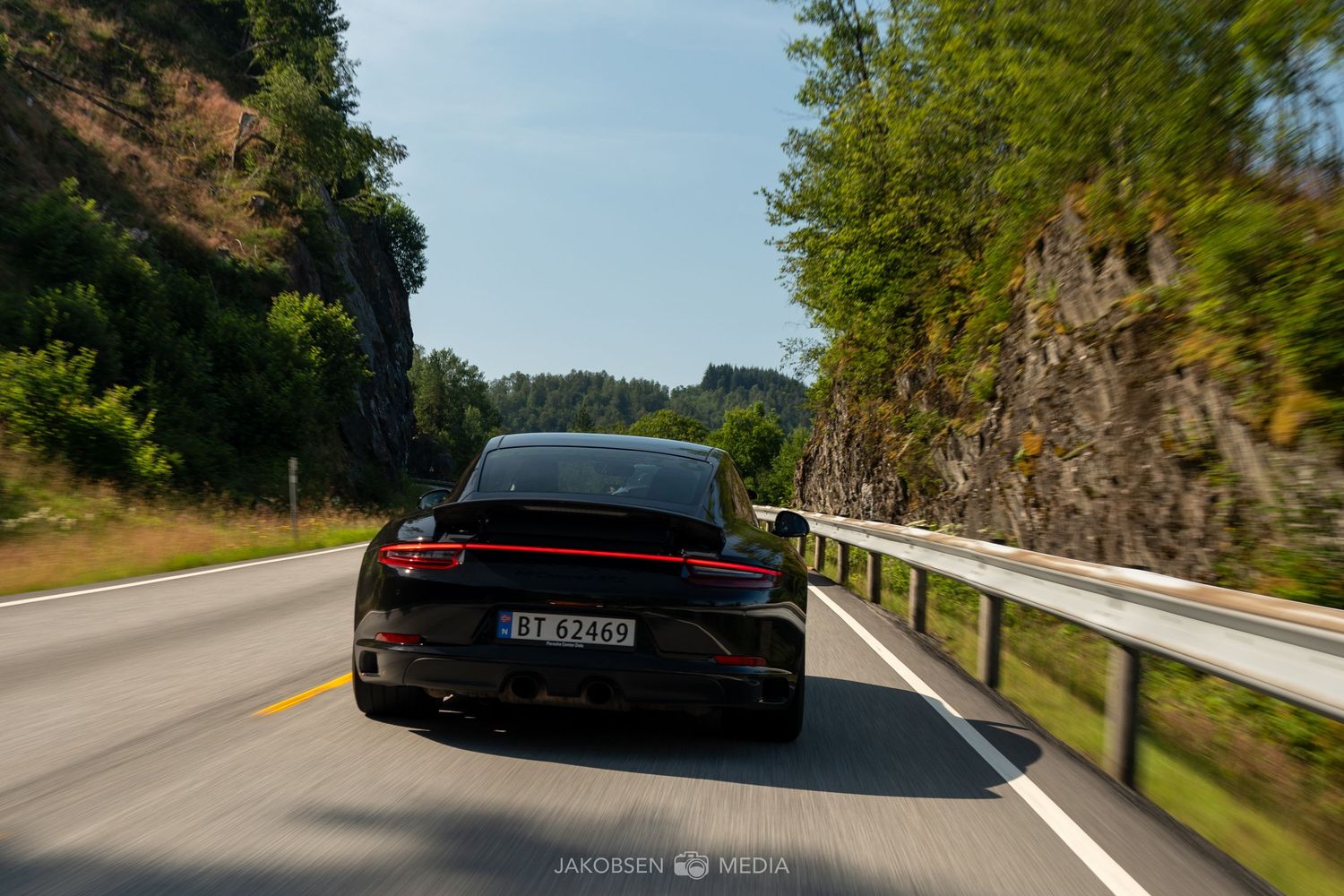 Rear view of a black Porsche driving on a scenic mountain road with rock walls.