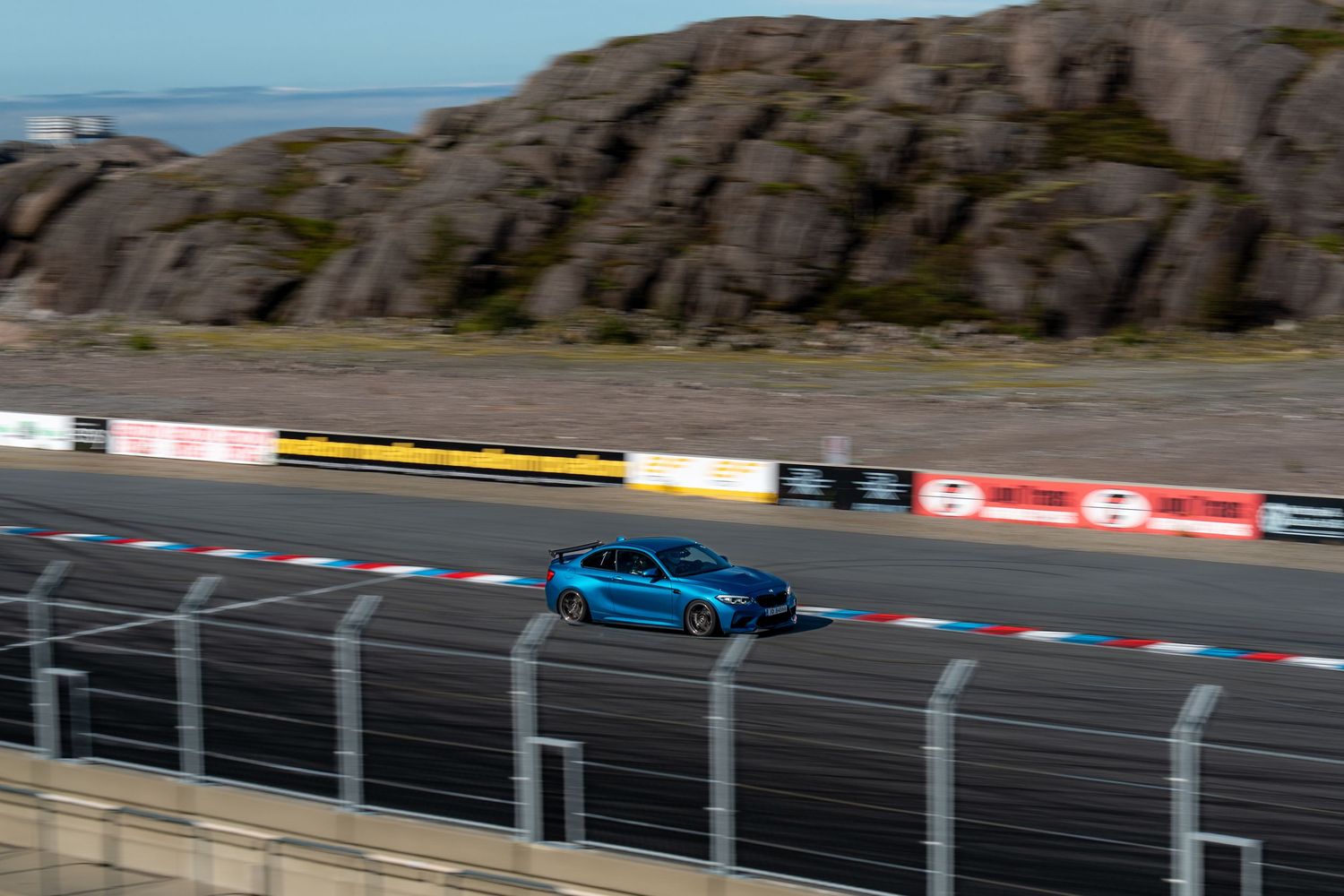 A blue sports car races around a curving racetrack with rocky cliffs in the background.