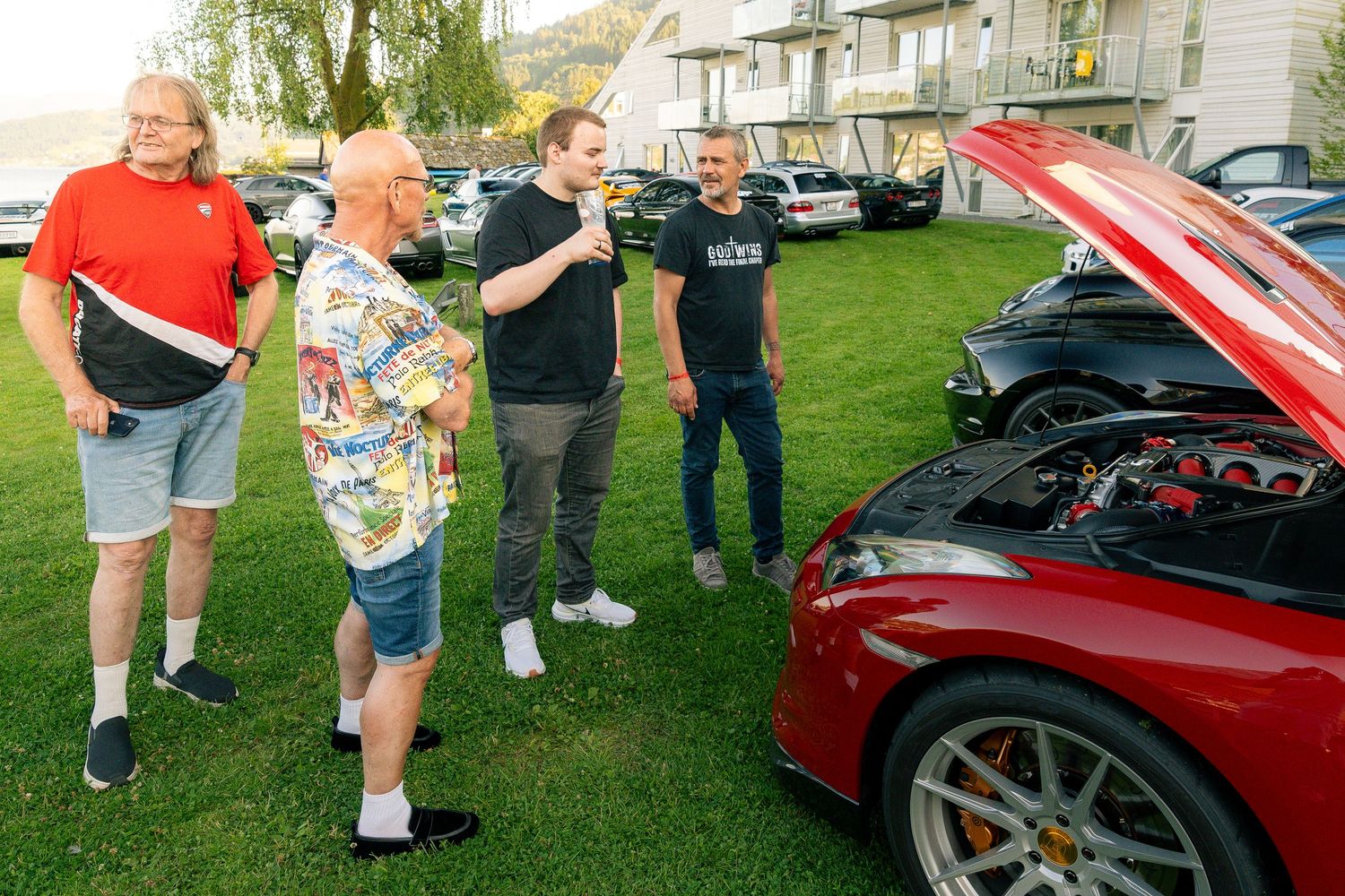 Four men chat beside a red Ferrari with its engine bay open at an outdoor car show on a grassy lawn.