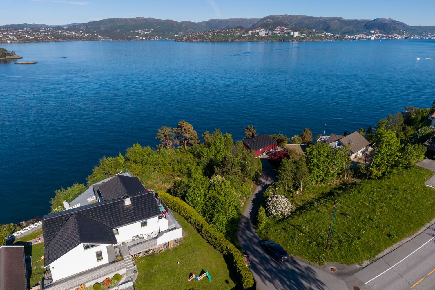 Aerial view of a white house with a dark roof overlooking a calm blue fjord, with green hillsides, nearby homes and distant mountains.