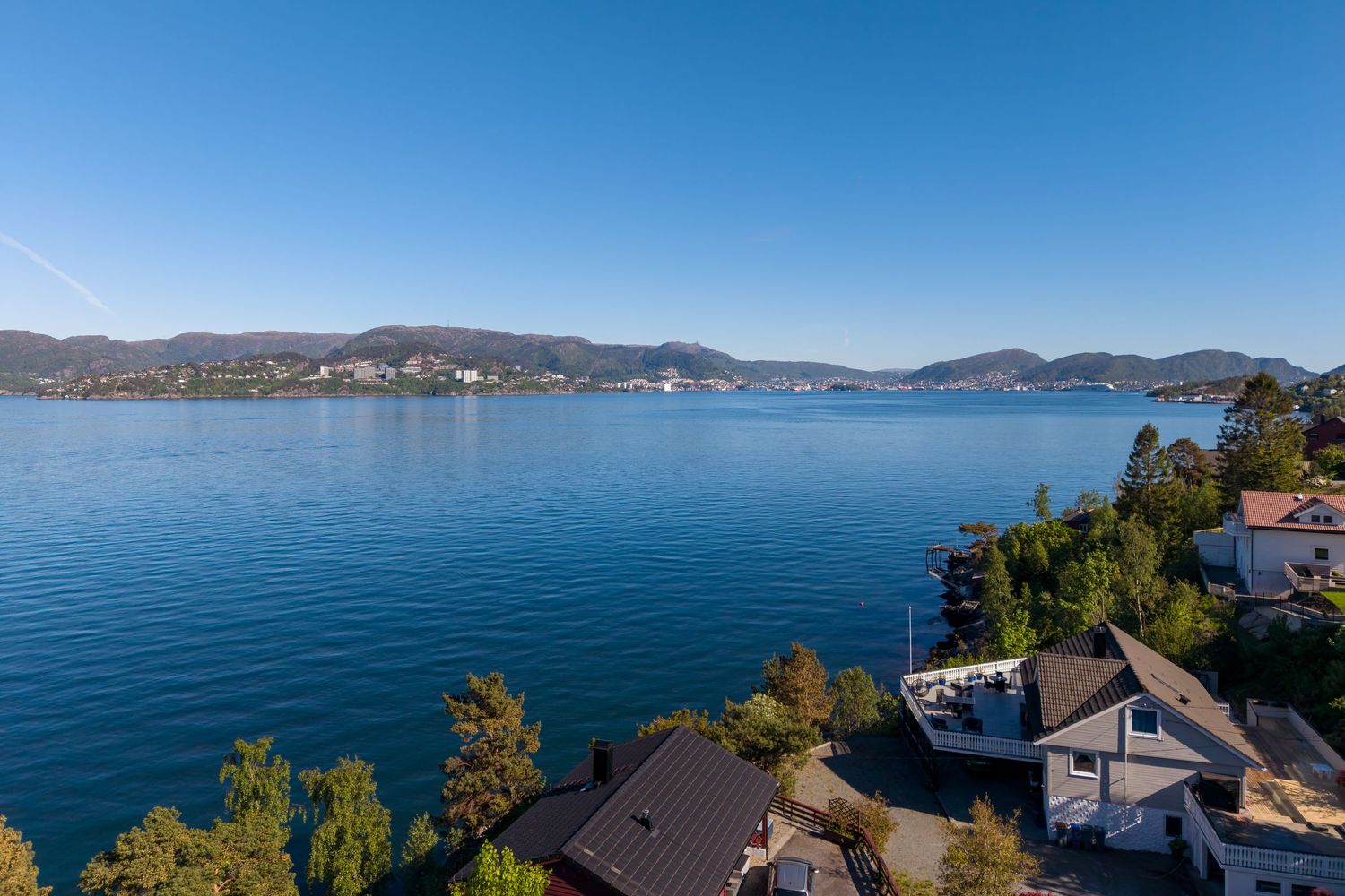Aerial view of waterfront homes and green trees along a calm blue fjord with distant mountains under a clear sky.