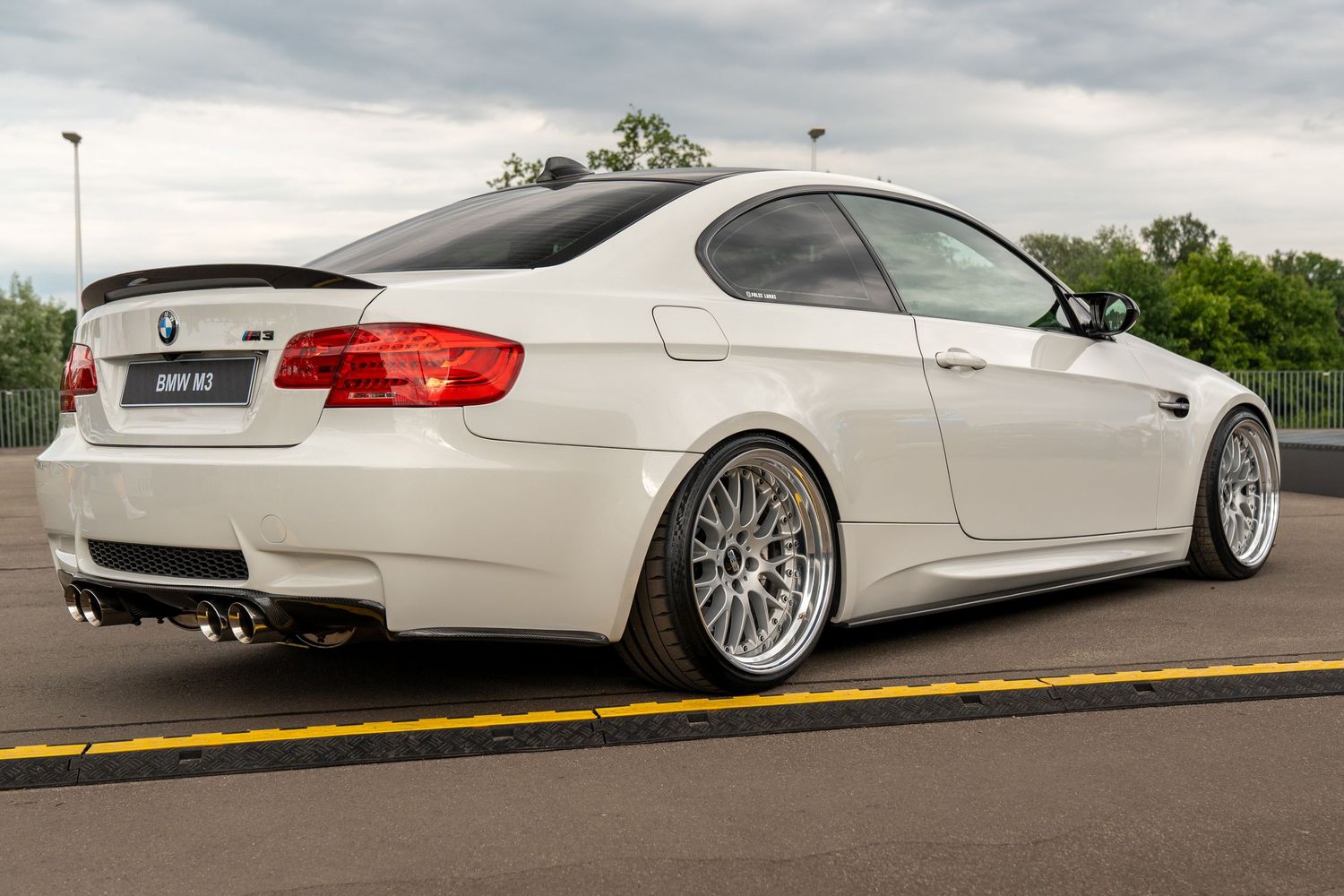 Side profile of a silver BMW M3 coupe with custom wheels and lowered suspension on a cloudy day.