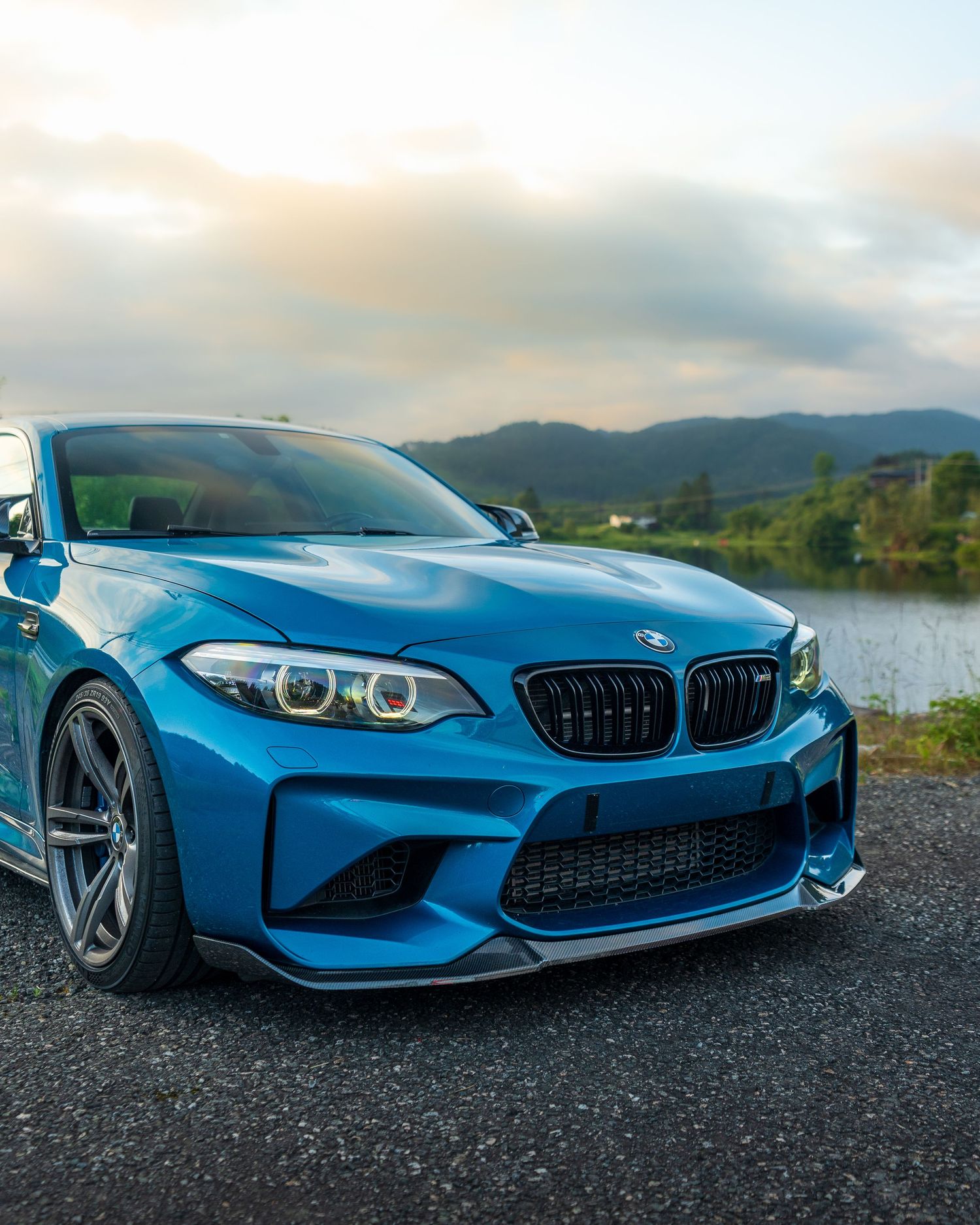 A sleek blue BMW M2 sports car photographed at sunset against mountains and a lake.