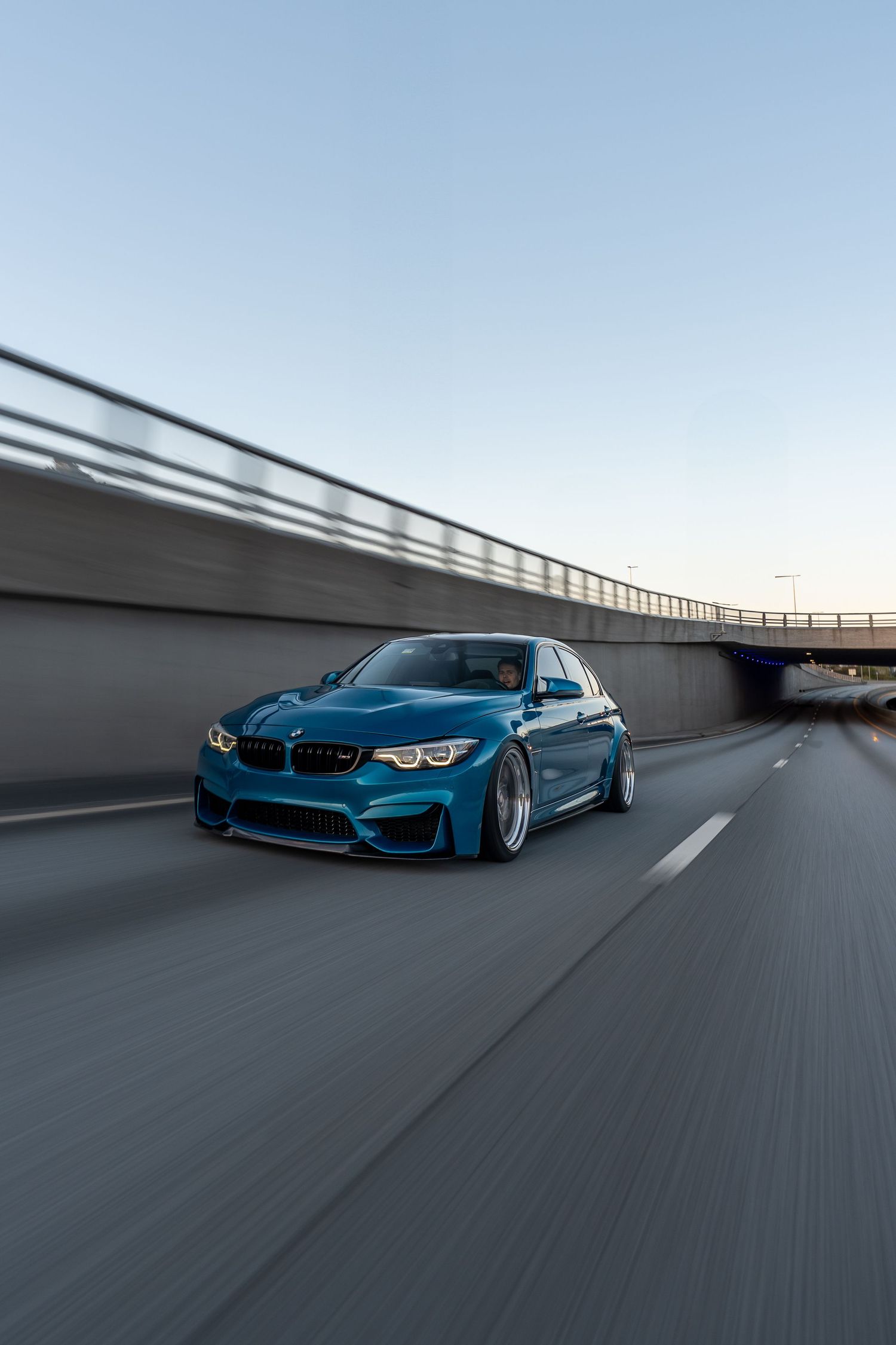 Lowered blue BMW M3 driving under bridge with motion blur effect.