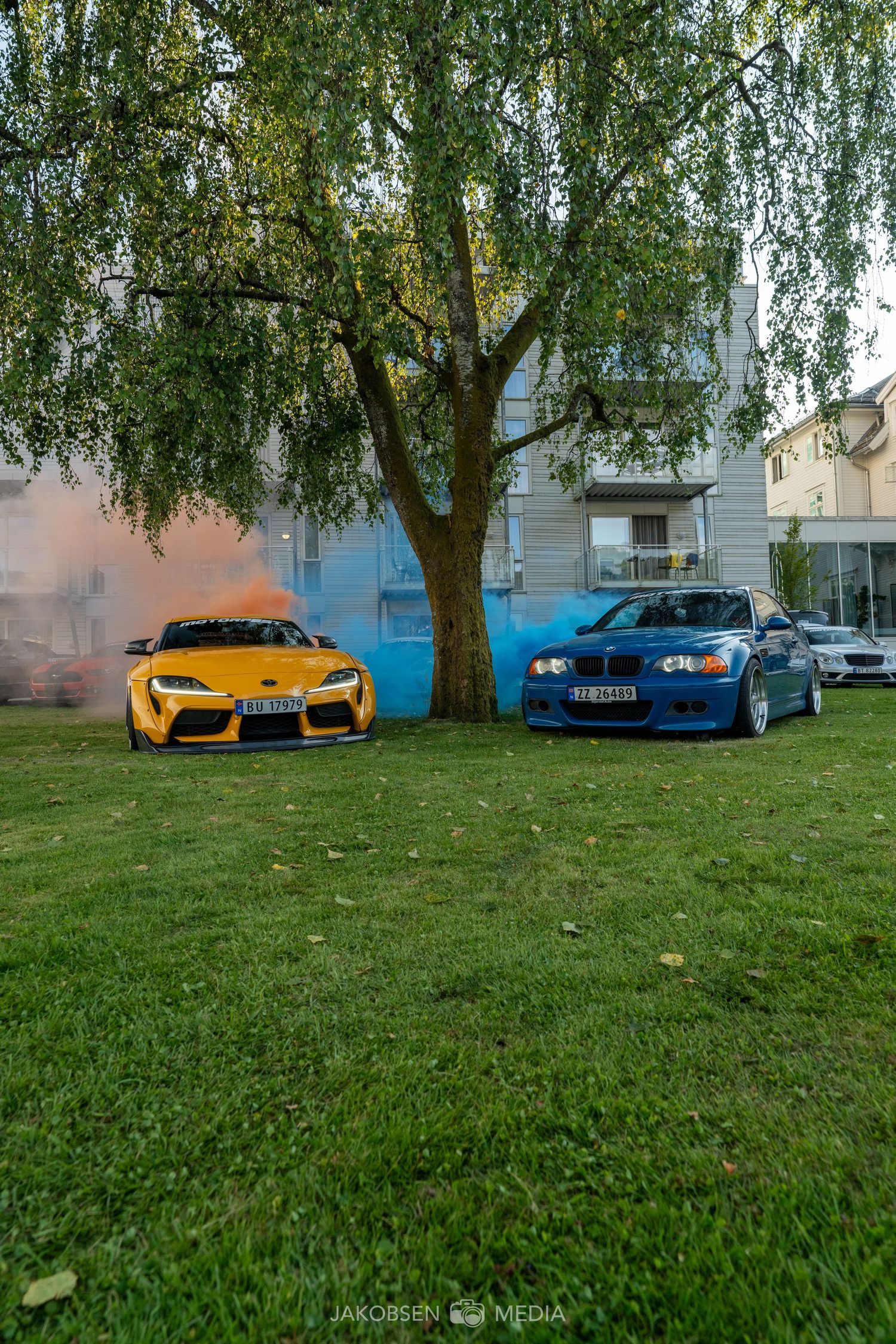 Vibrant orange and blue sports cars parked on grass against a backdrop of birch trees.