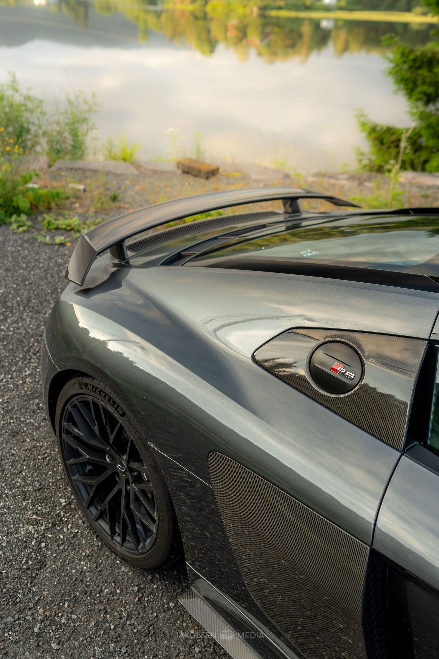 Close-up view of a gray car's front fender and wheel with black multi-spoke rims parked near a misty lake.