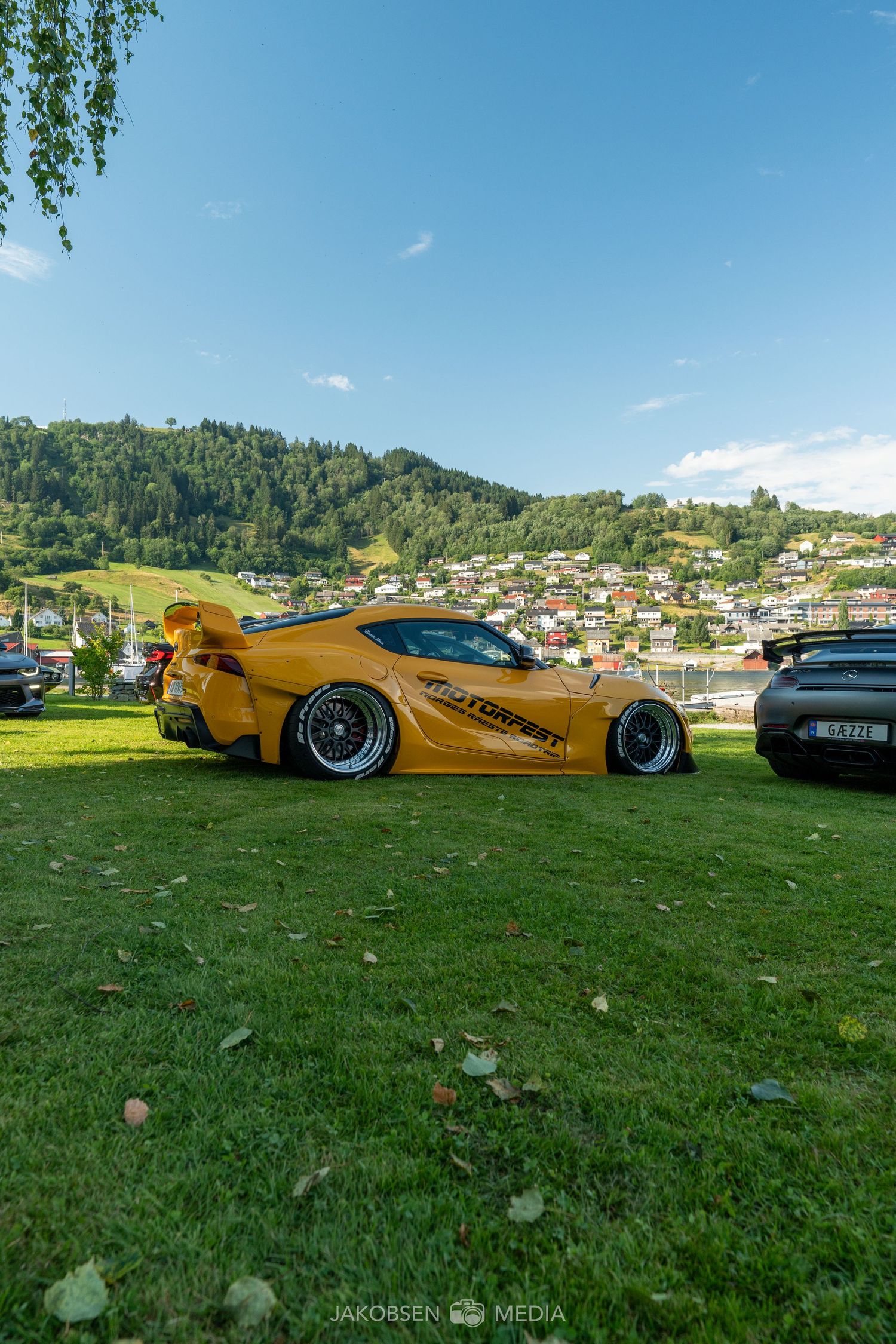 Yellow sports car displayed on green grass with mountain landscape and houses in the background.