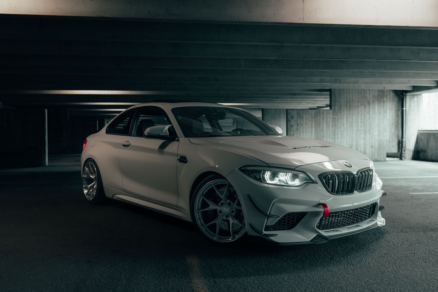 A sleek BMW M2 sports car in matte gray finish is parked in a dimly lit underground parking garage.
