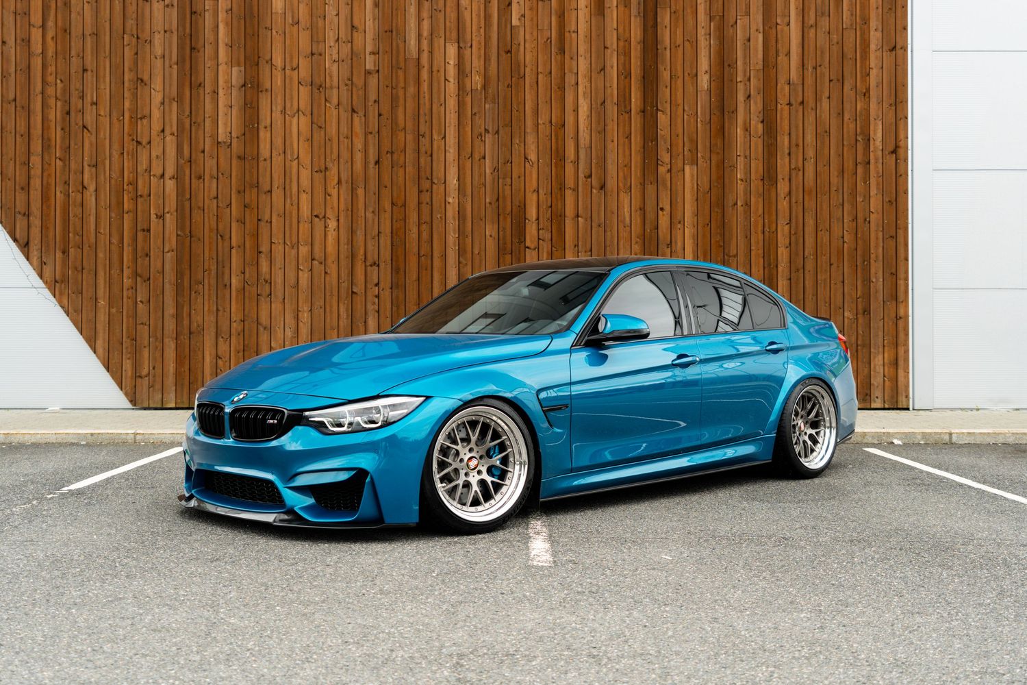 A modified BMW M3 F80 in metallic blue sits lowered on custom bronze wheels against a wooden wall background.