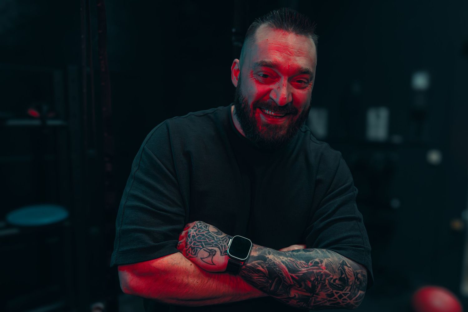 Tattooed person wearing dark clothing photographed with moody red lighting.