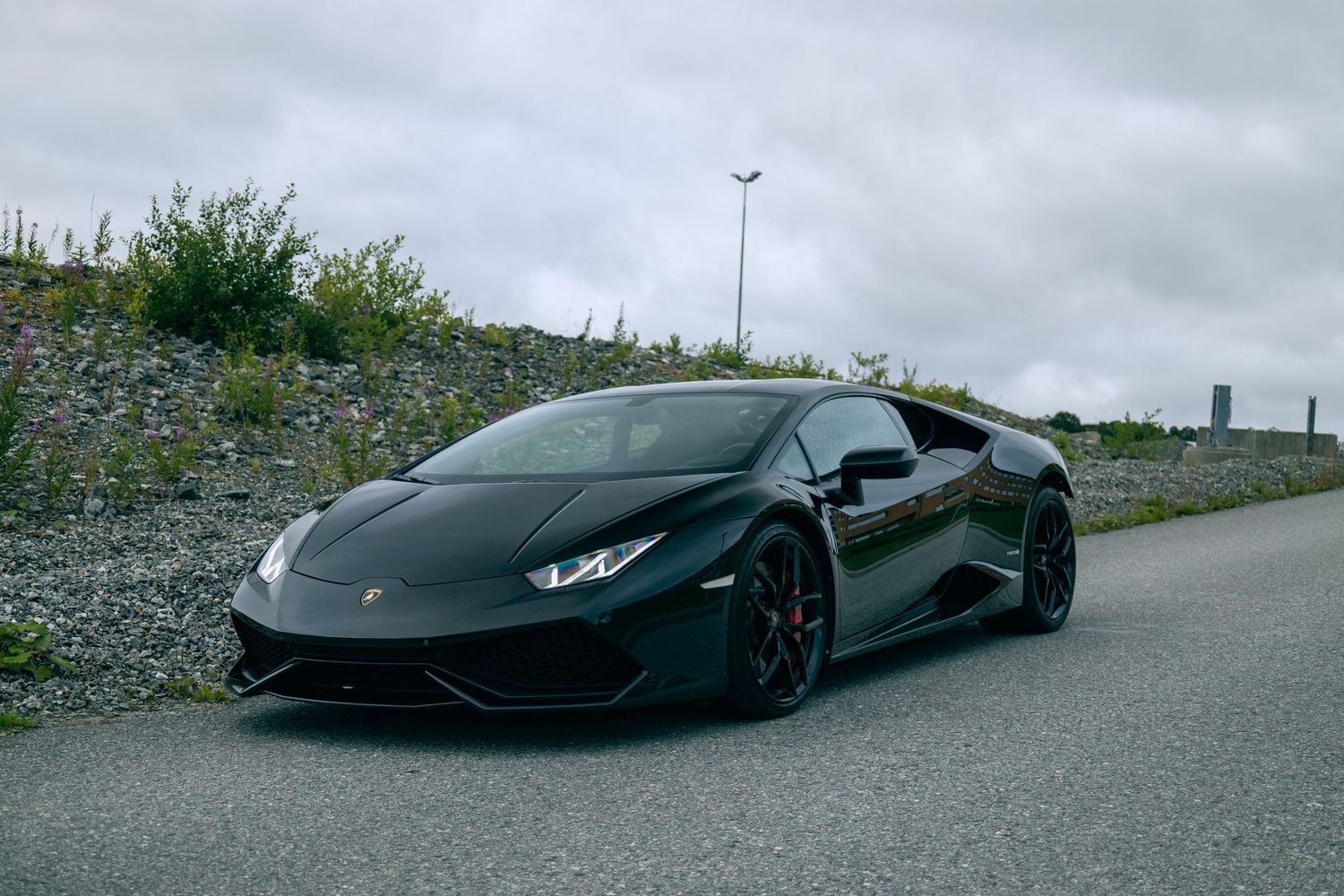 Matte black Lamborghini Huracan parked on gravel lot under cloudy skies.