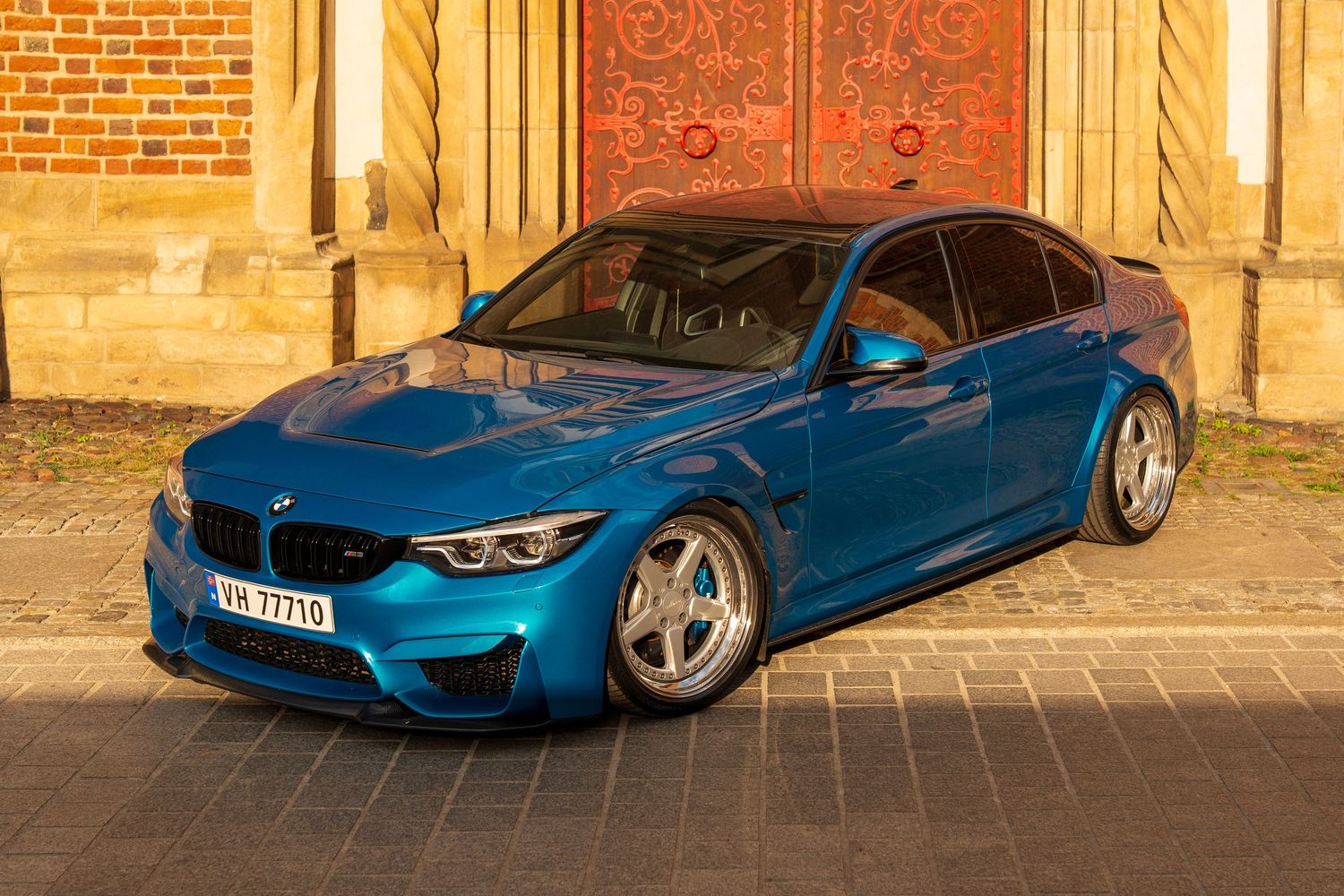 Modified BMW M3 in metallic blue parked against a brick and stone building facade.
