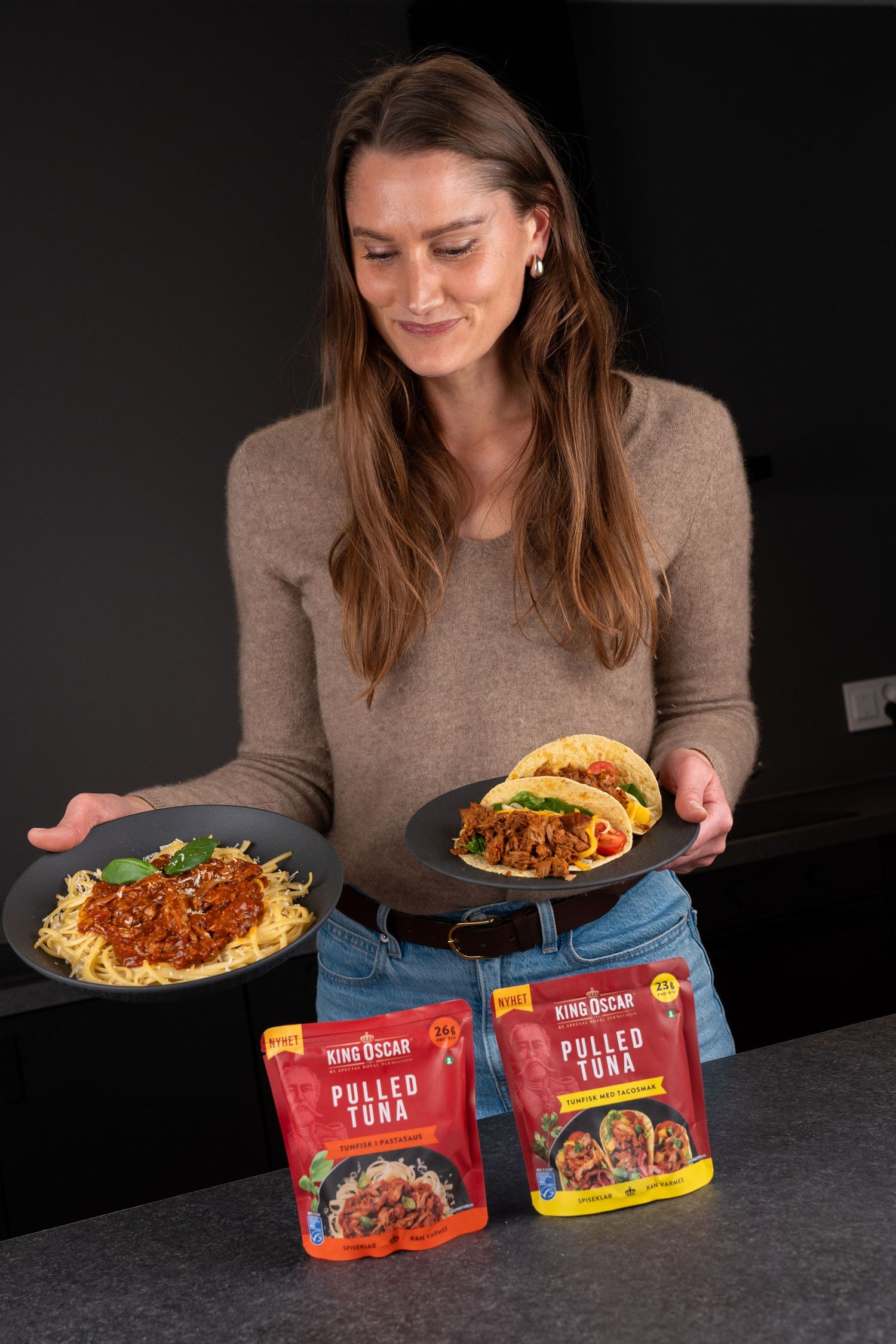 Woman holding plates of spaghetti and tacos made with King Oscar Pulled Tuna pouches on a kitchen counter.