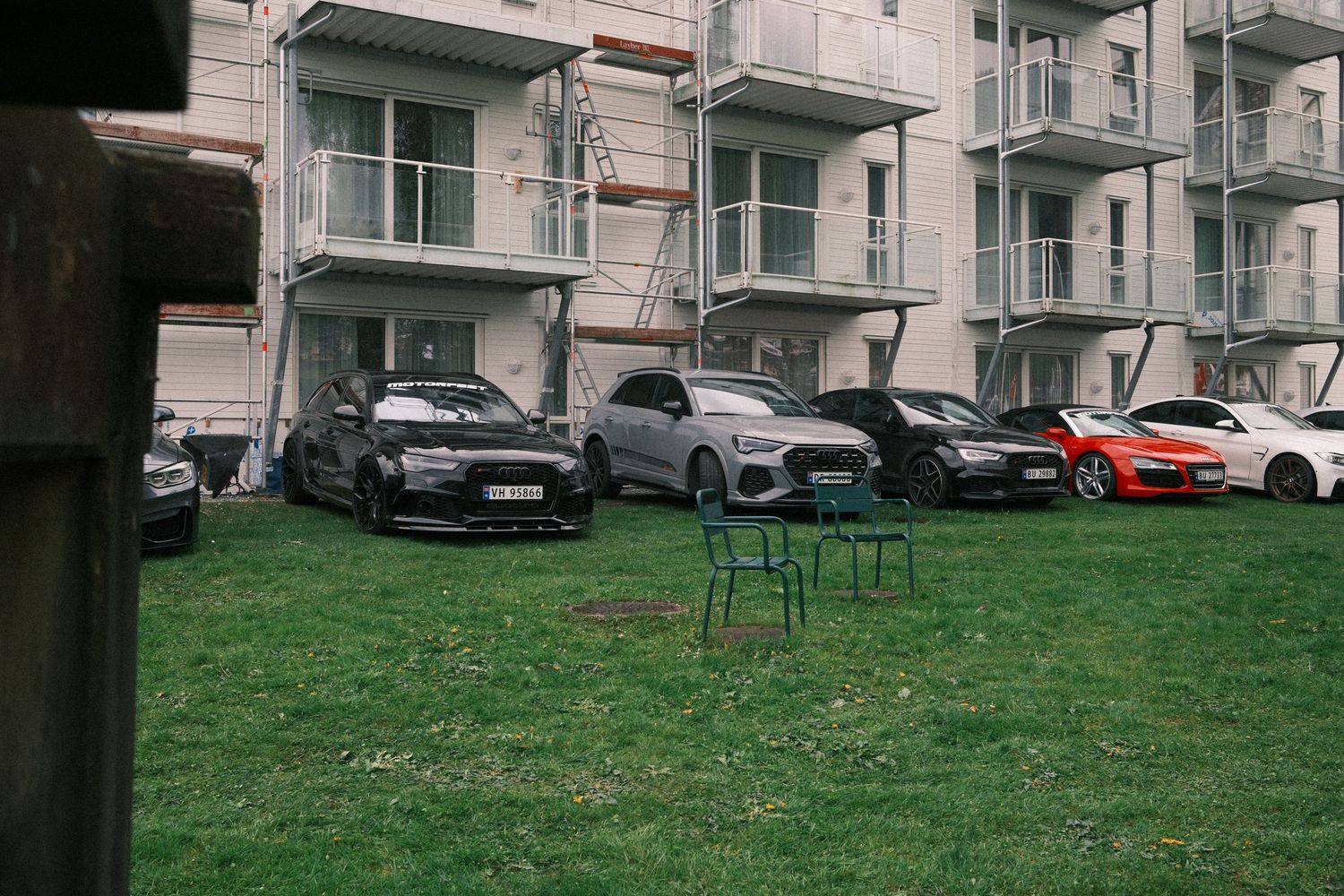 Row of parked sports cars on a grassy lawn beside an apartment building, with two green metal chairs in the foreground.
