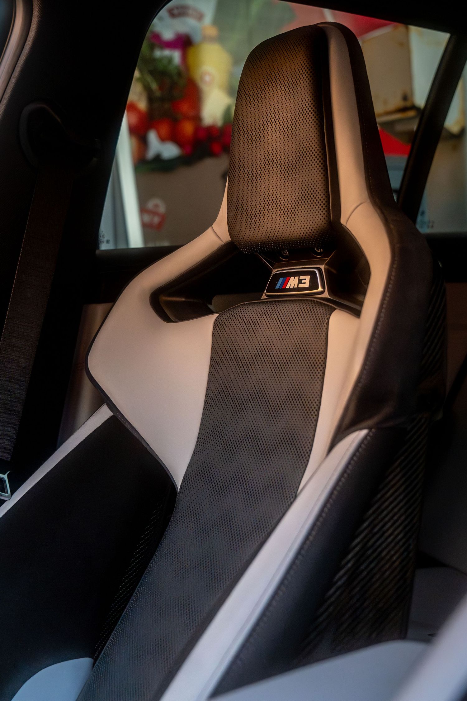 BMW M sport car interior featuring a black and white leather bucket seat with M logo badge and carbon fiber accents.