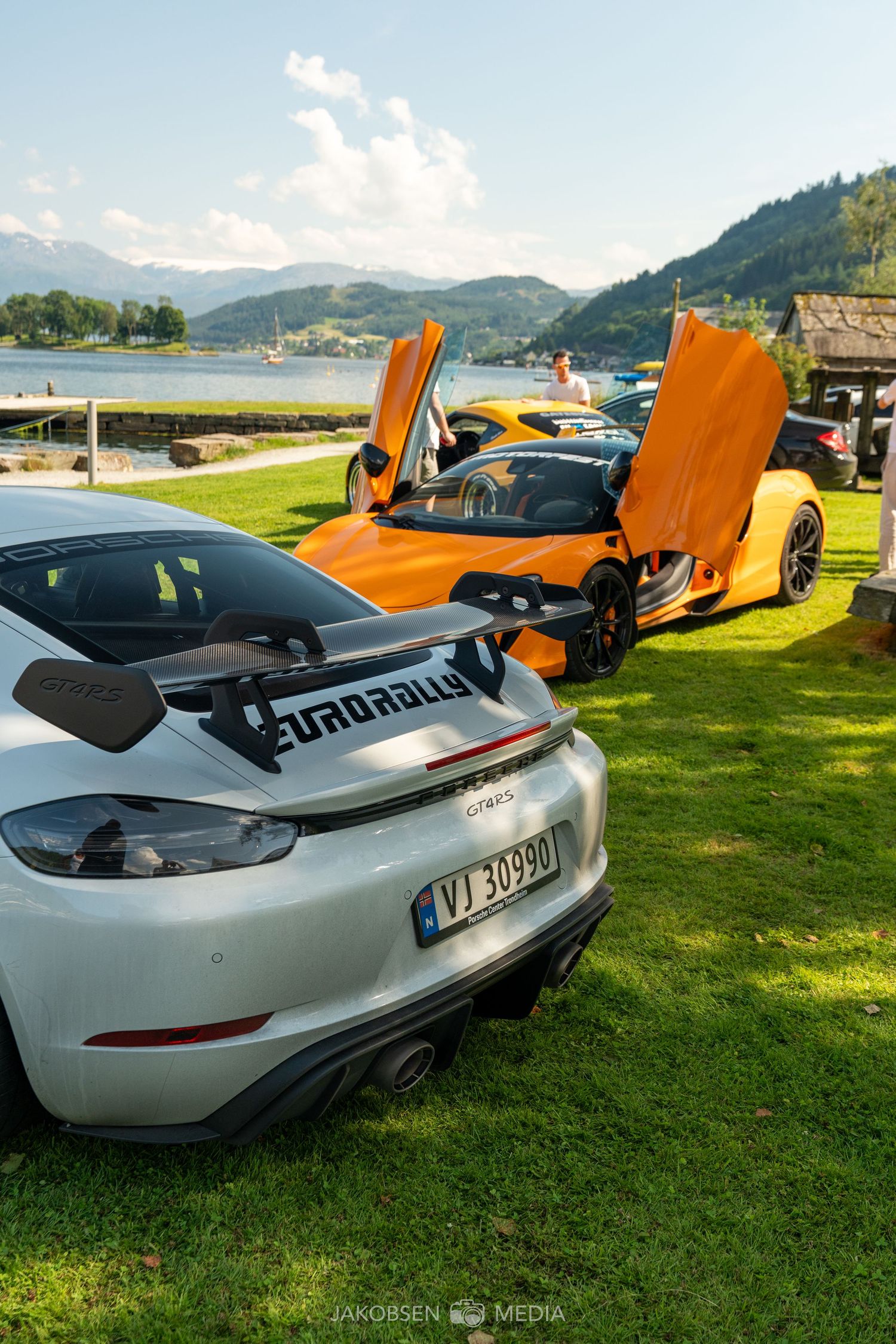 Row of exotic supercars including orange and white models displayed on a lakeside lawn.