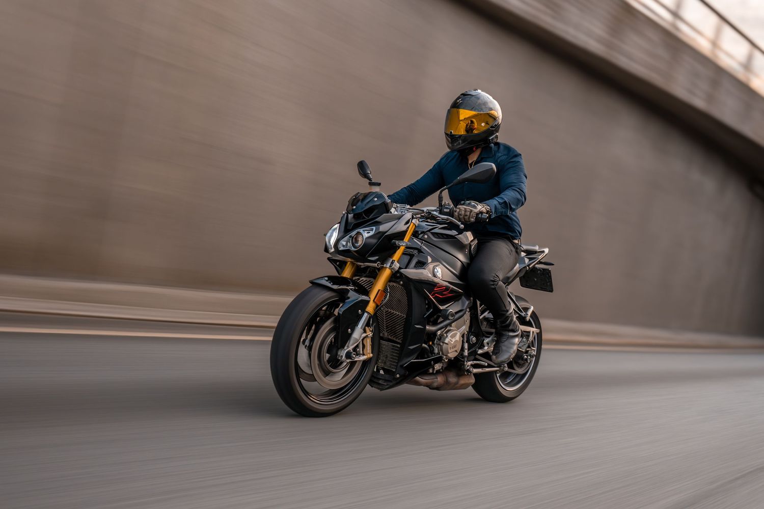 Motorcyclist in blue winter gear rides a black and gold sport bike at speed through an urban tunnel.