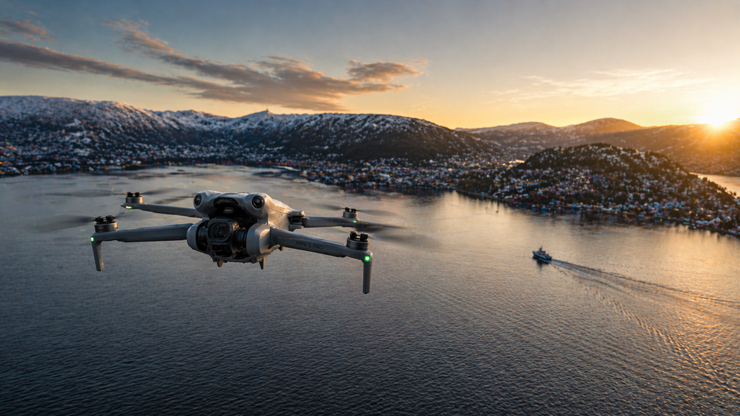A DJI drone flies over a scenic Norwegian fjord at sunset with snow-capped mountains and a coastal town in the background.