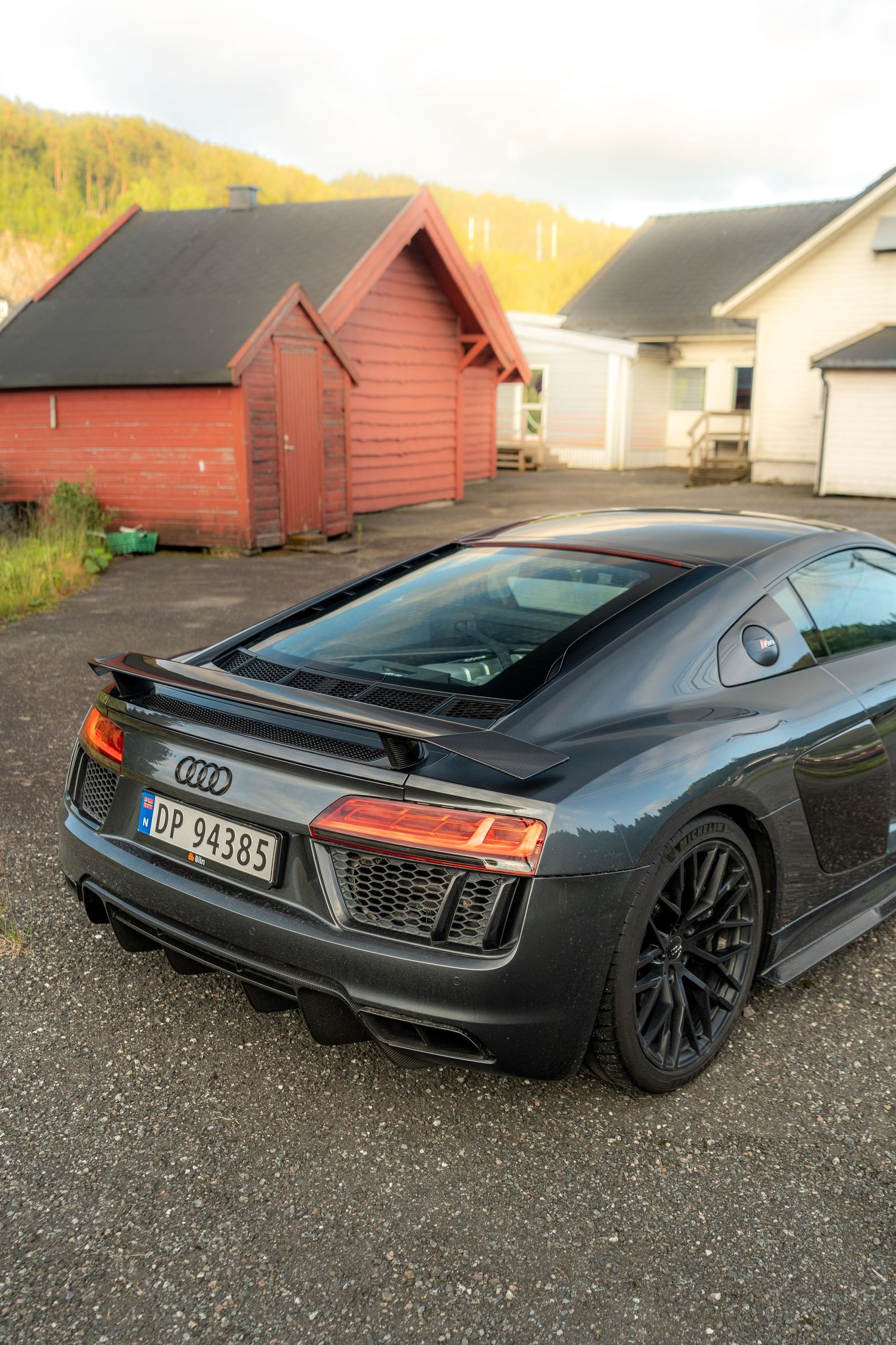 A sleek black Audi R8 sports car parked in front of red wooden buildings at dusk.