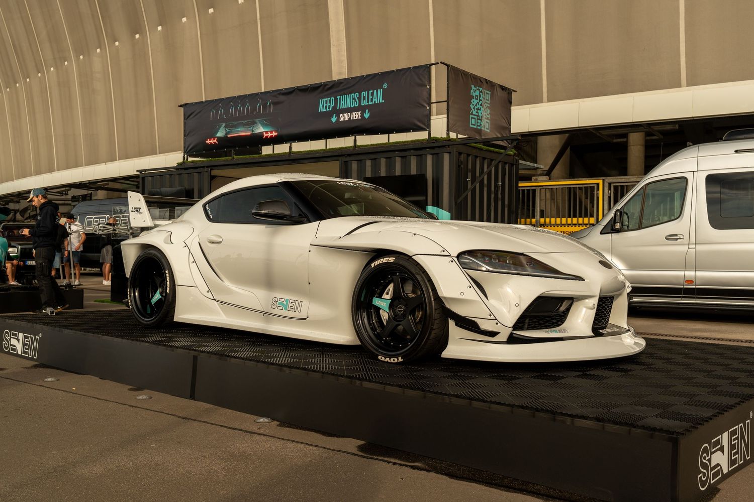 A modified white Toyota Supra with wide body kit and low profile stance sits on display at a car show.