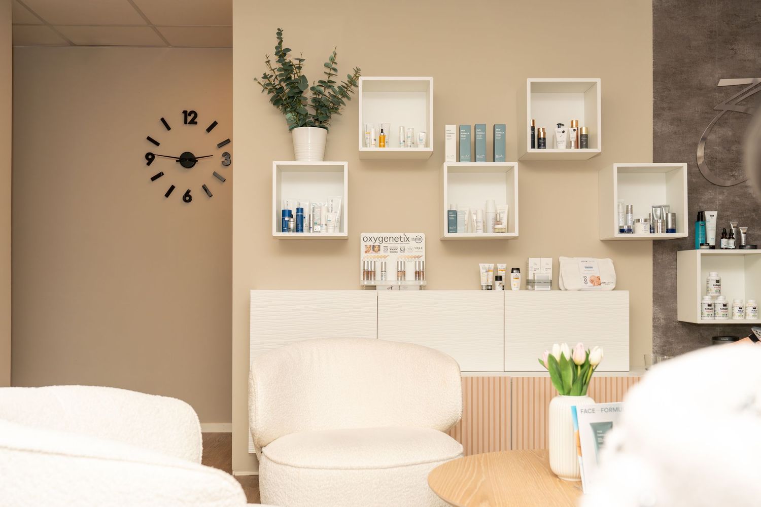 Modern beauty salon waiting area with cream armchairs, wall-mounted cube shelves displaying skincare products, a white sideboard, tulips and