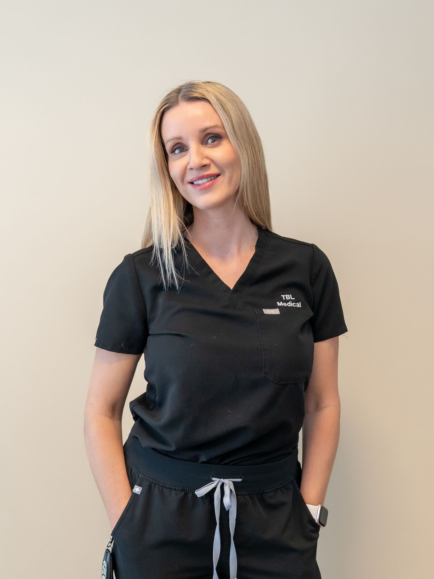Smiling woman with long blonde hair wearing black medical scrubs with a logo, standing with her hands in her pockets.