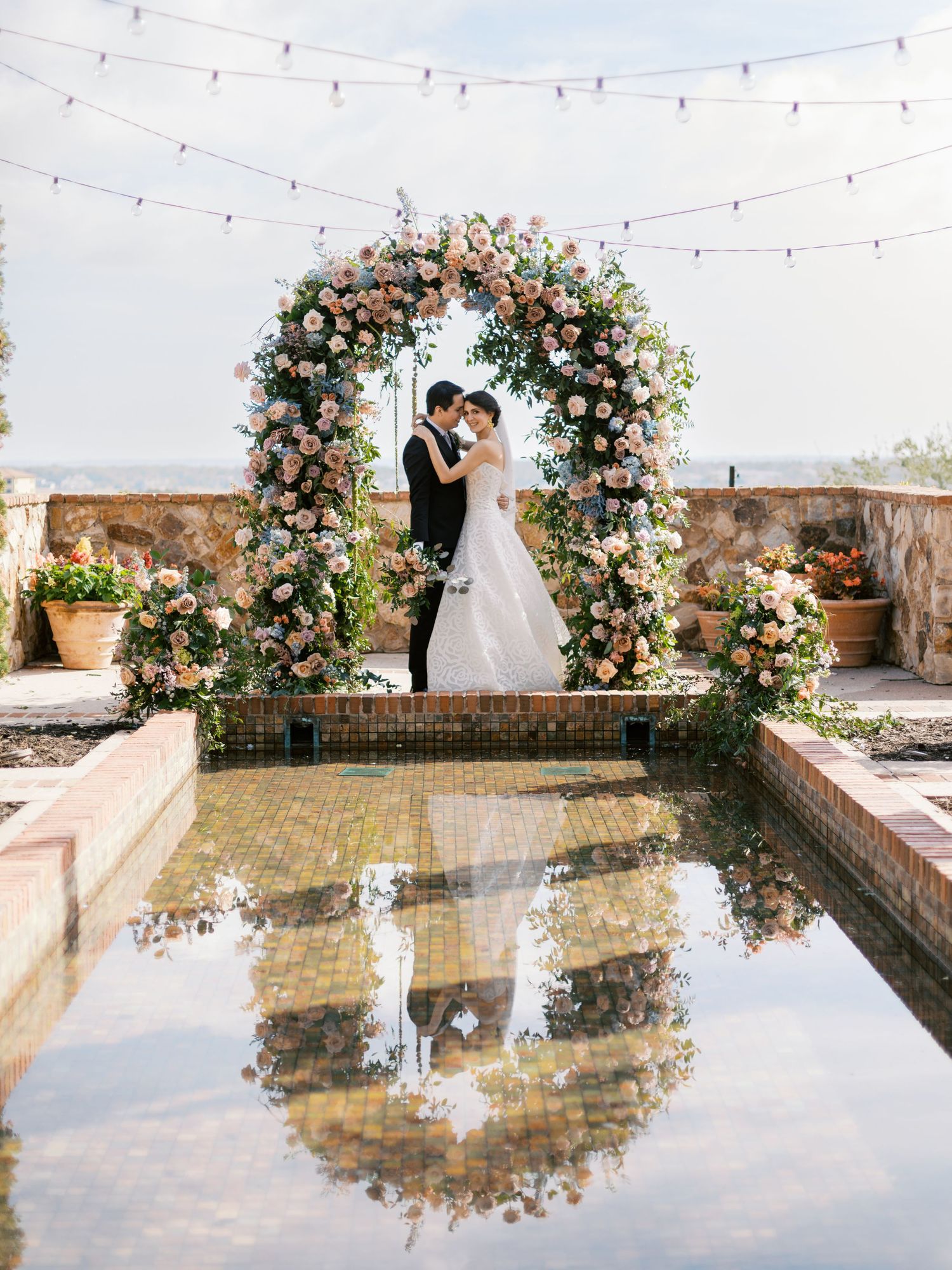 A romantic wedding arch covered in pink and white flowers stands over a reflecting pool with string lights overhead.