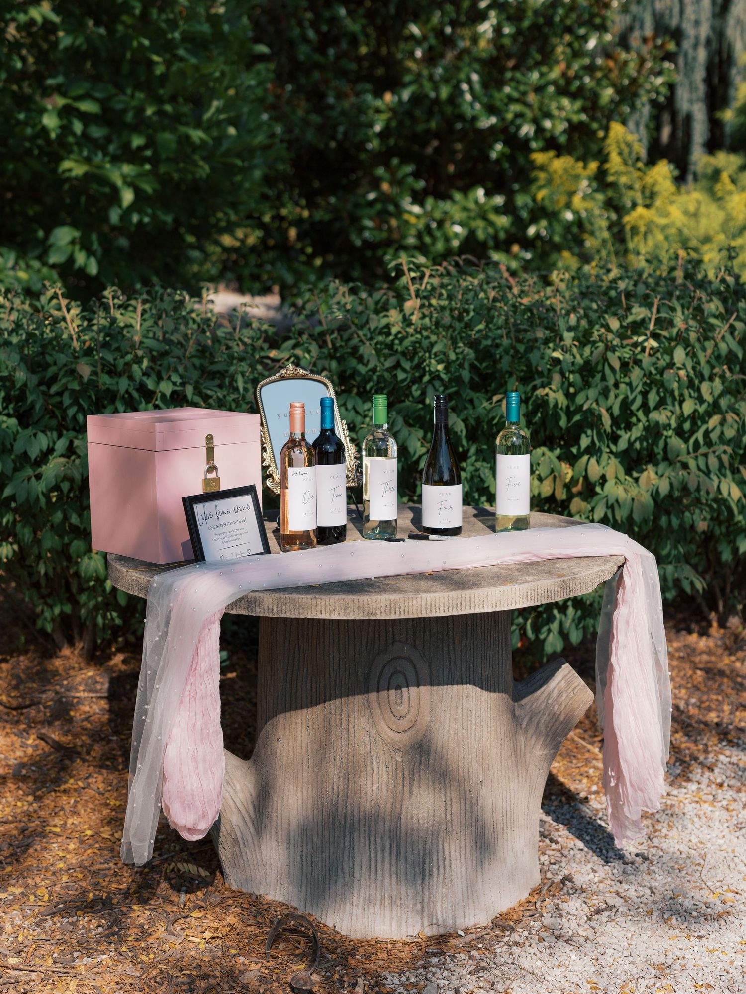 A rustic wooden table displays wine bottles and decor against a garden backdrop.