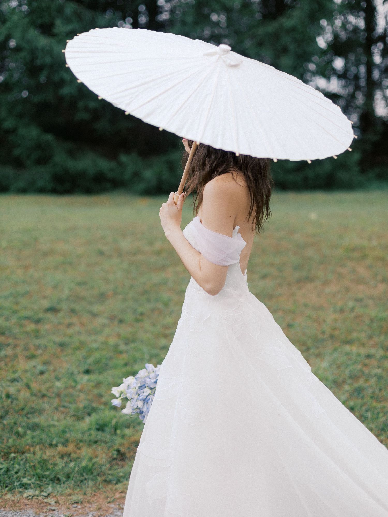 Bride in flowing white dress holding a white parasol in a garden setting.