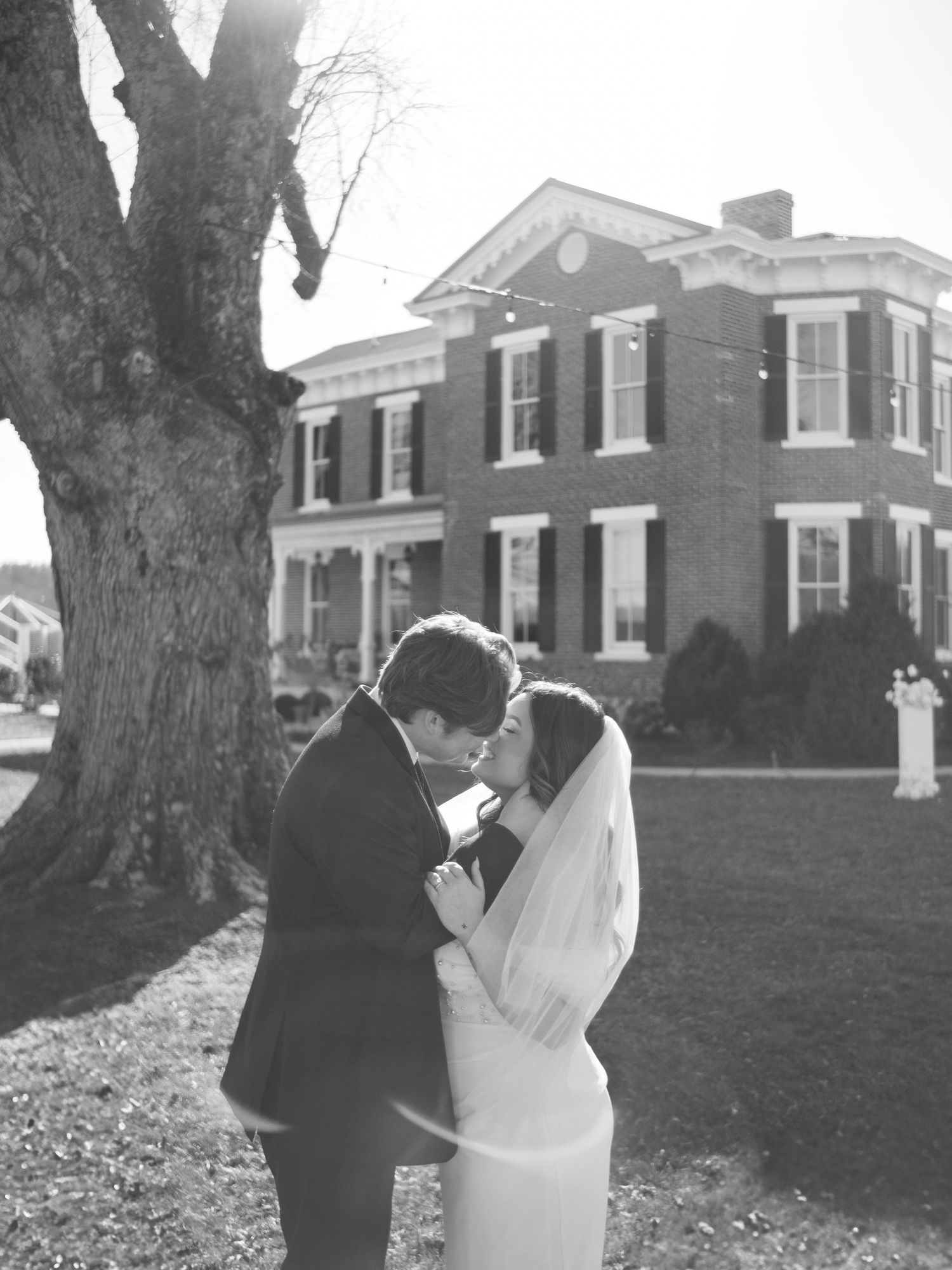 Black and white photo of newlyweds sharing an intimate moment in front of historic brick mansion.