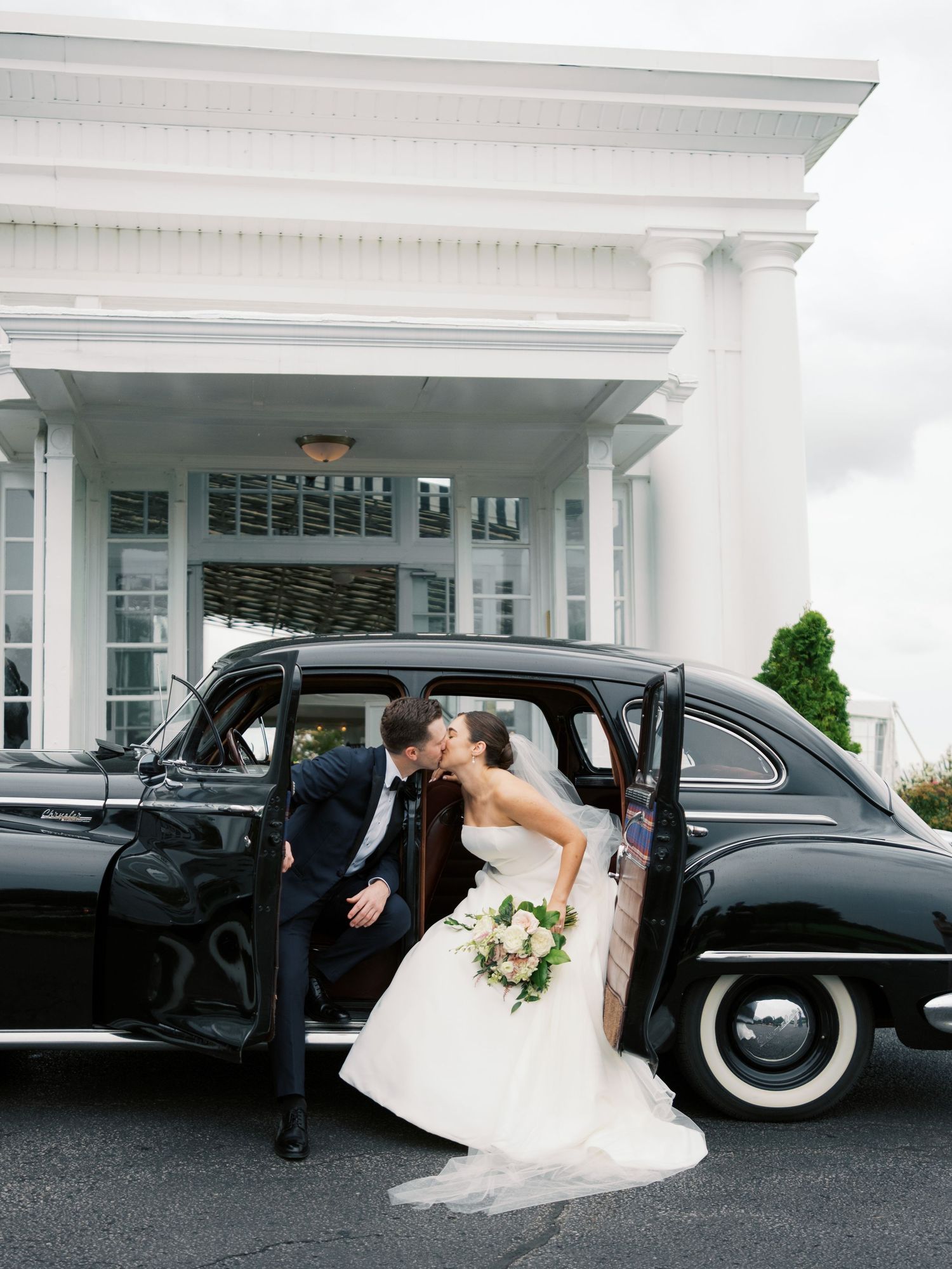 Classic black vintage car parked outside white pillared venue on wedding day.
