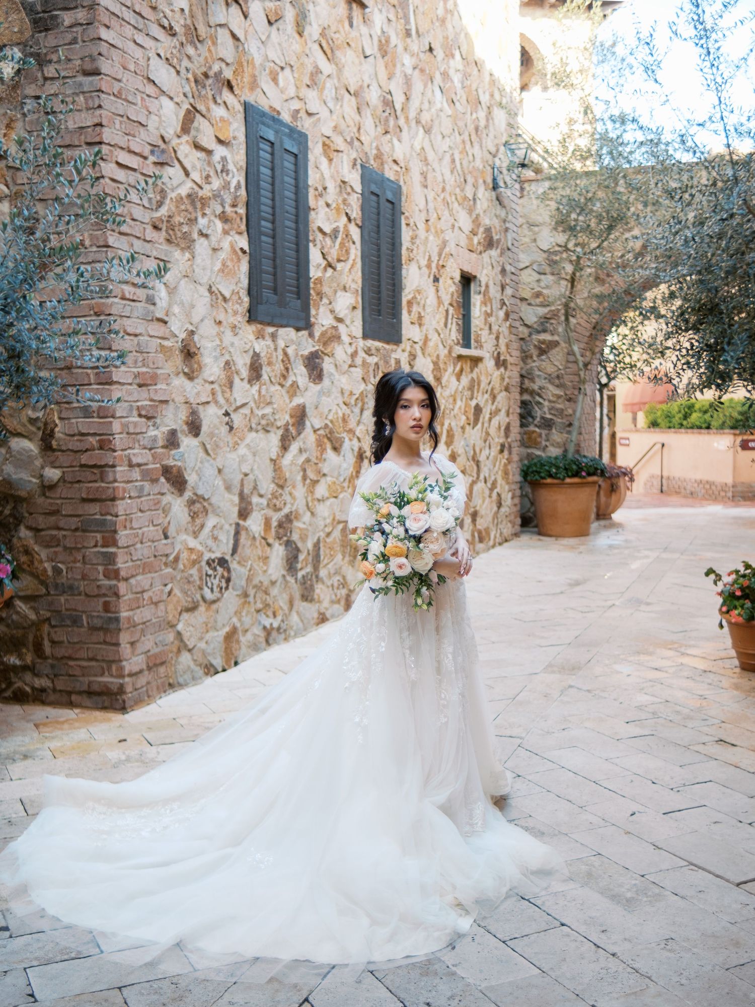 Bride in flowing white gown with long train poses against European stone wall.
