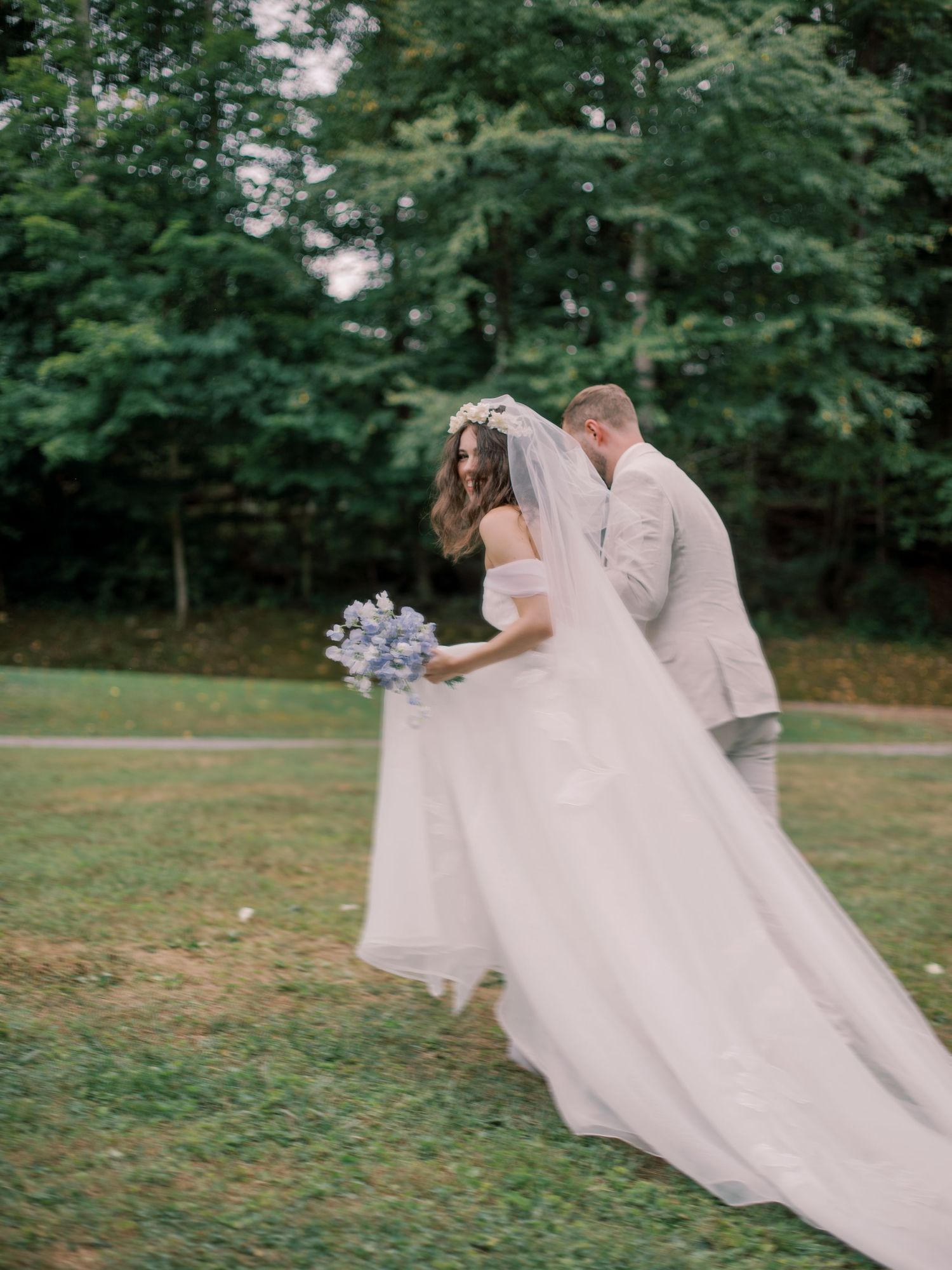 A couple in wedding attire walking across a grassy lawn holding a blue bouquet.