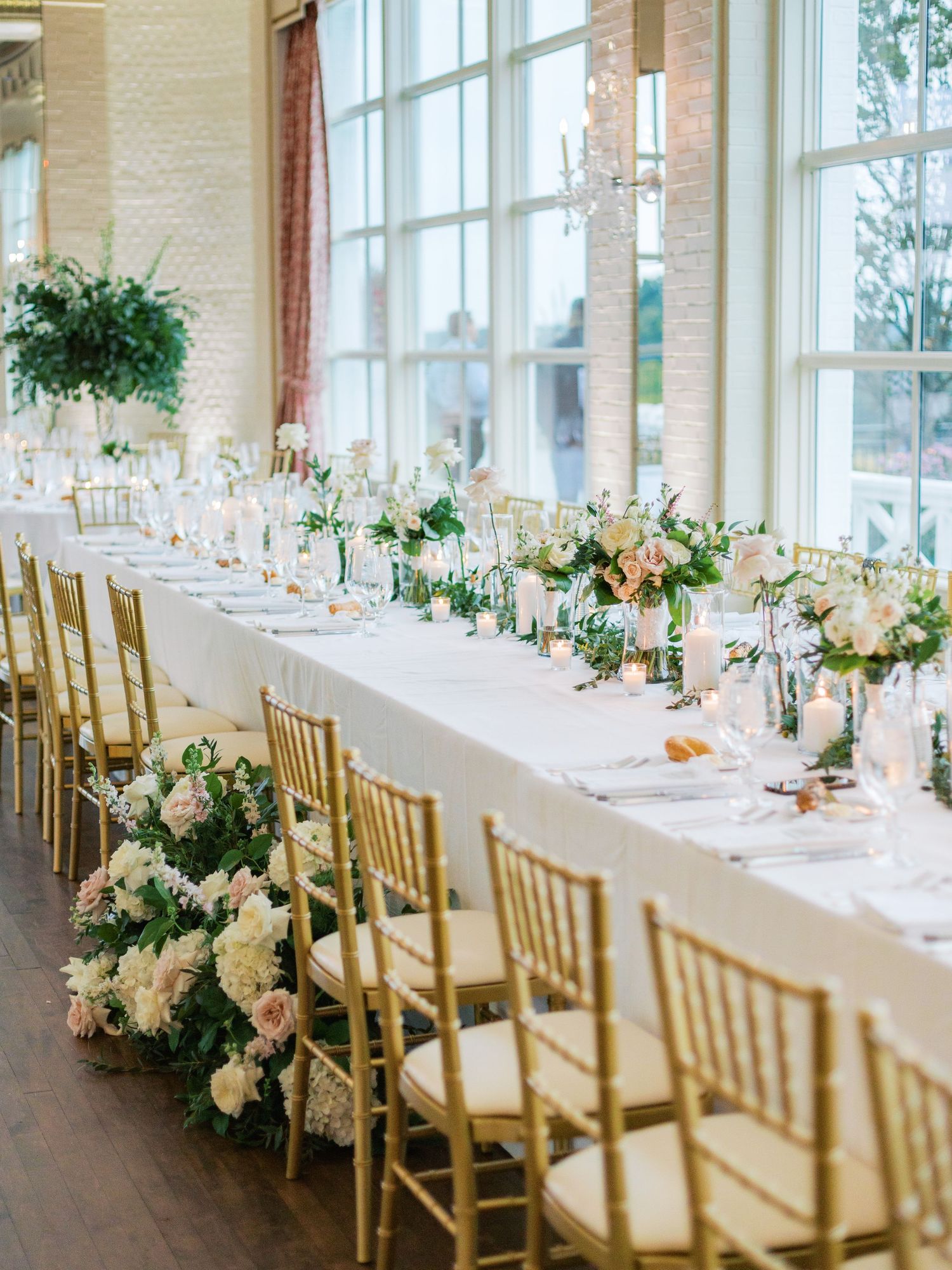Elegant wedding reception table with white linens, gold chairs, and lush floral centerpieces near tall windows.