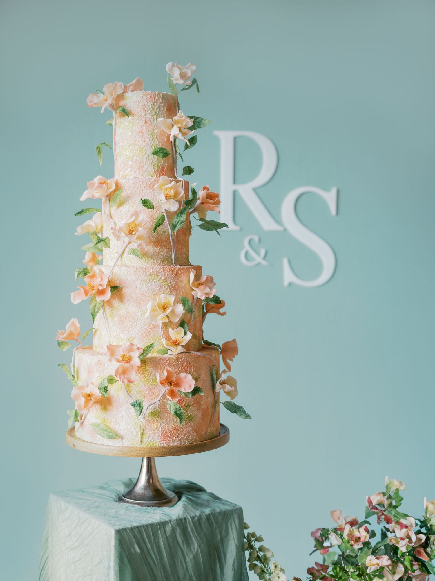 Elegant tiered wedding cake with coral sugar flowers and monogram against turquoise background.