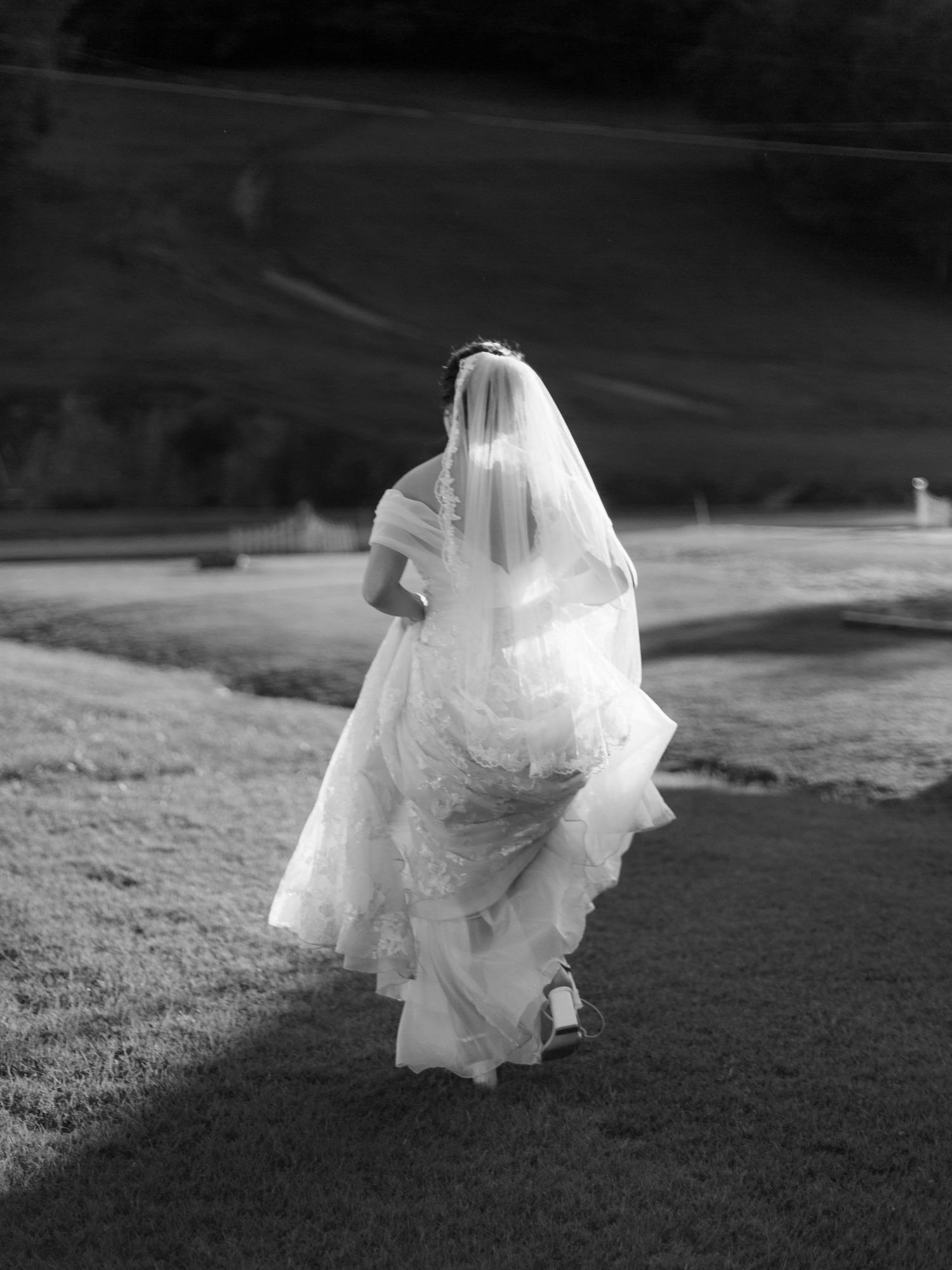 Black and white photo of a bride in flowing dress walking across a grassy field.