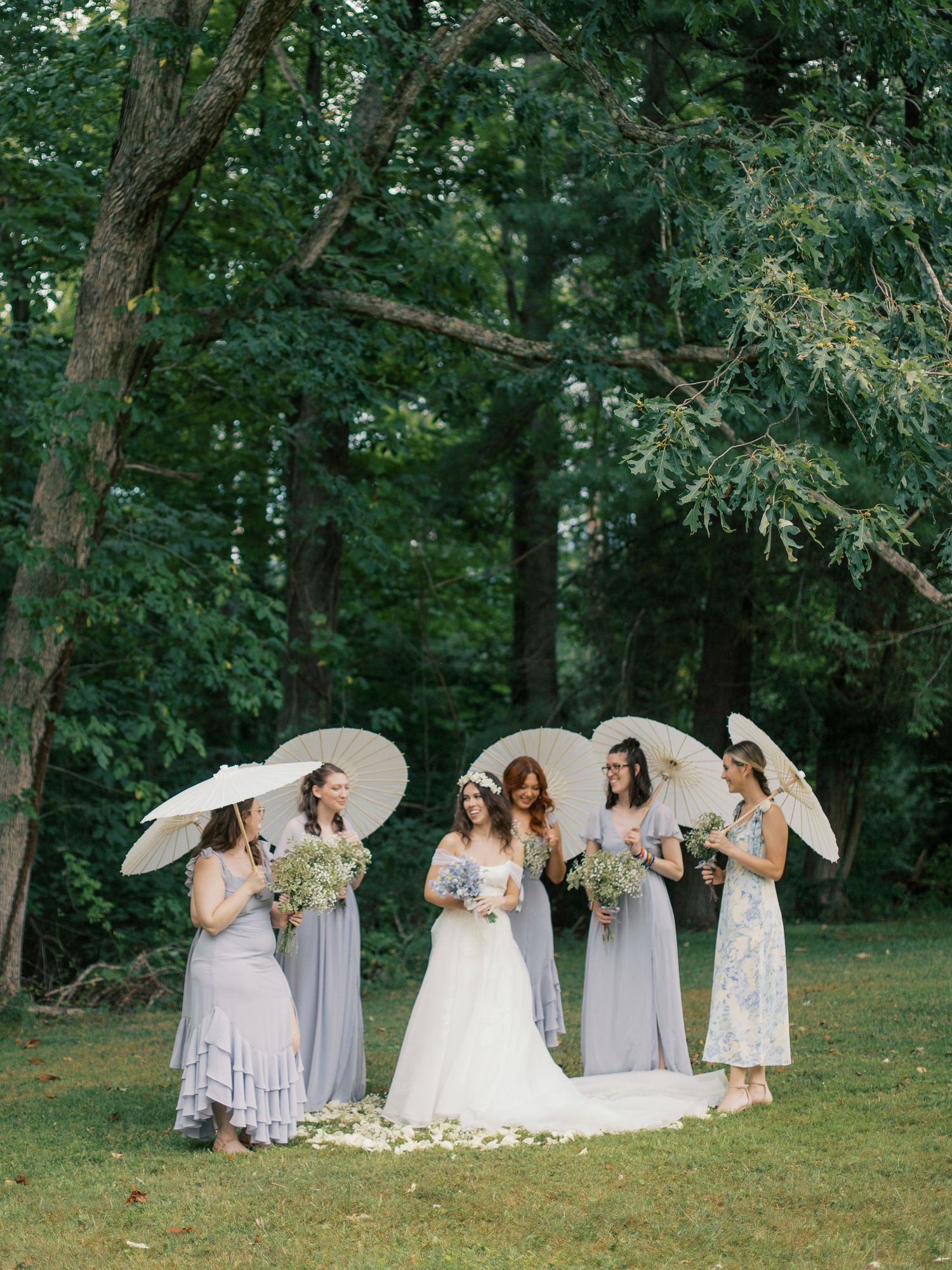 Bridal party wearing light colored dresses pose with white parasols in a lush garden setting.