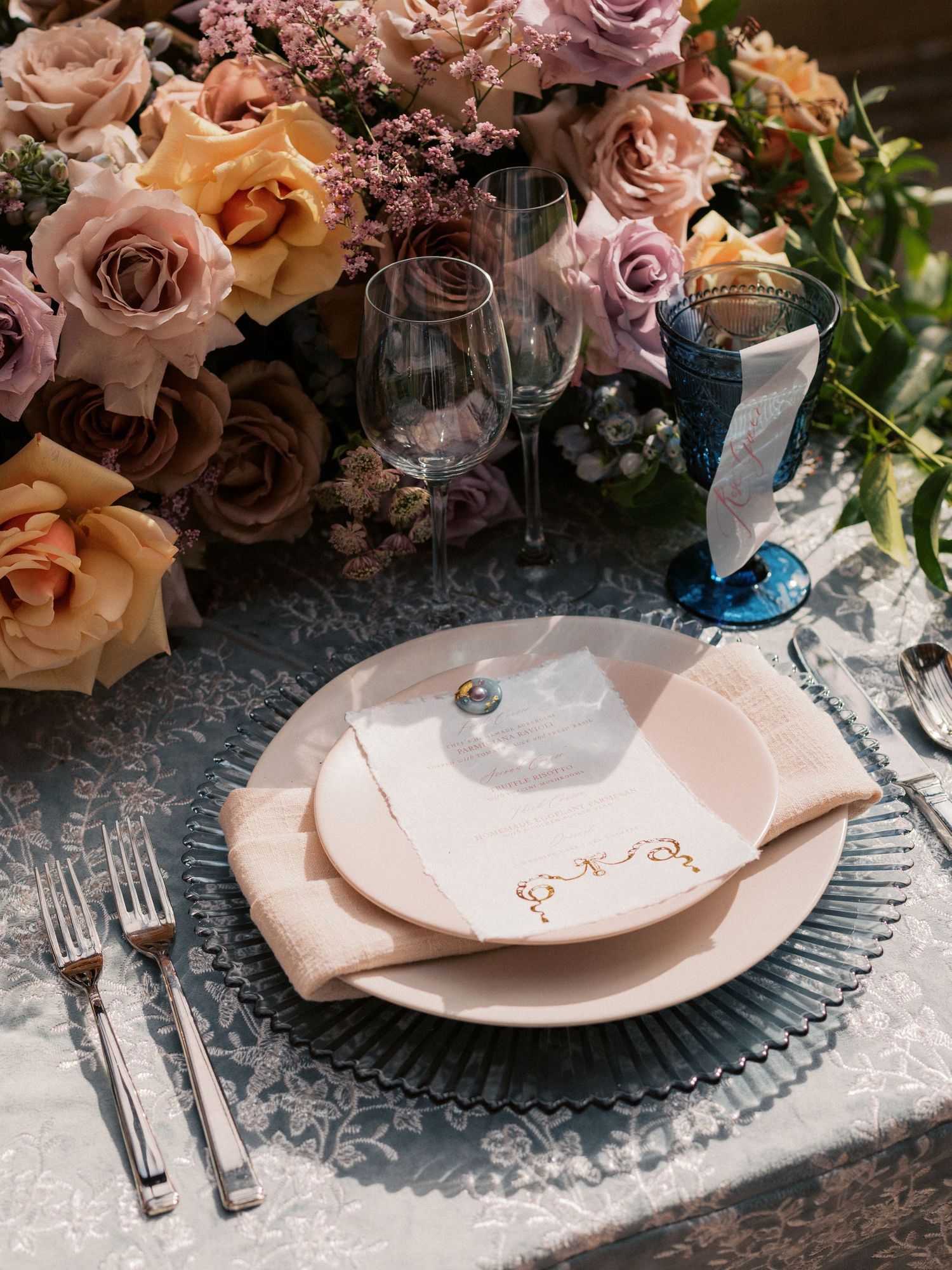 Romantic place setting with pink roses, wine glasses, and blush plates on lace tablecloth.