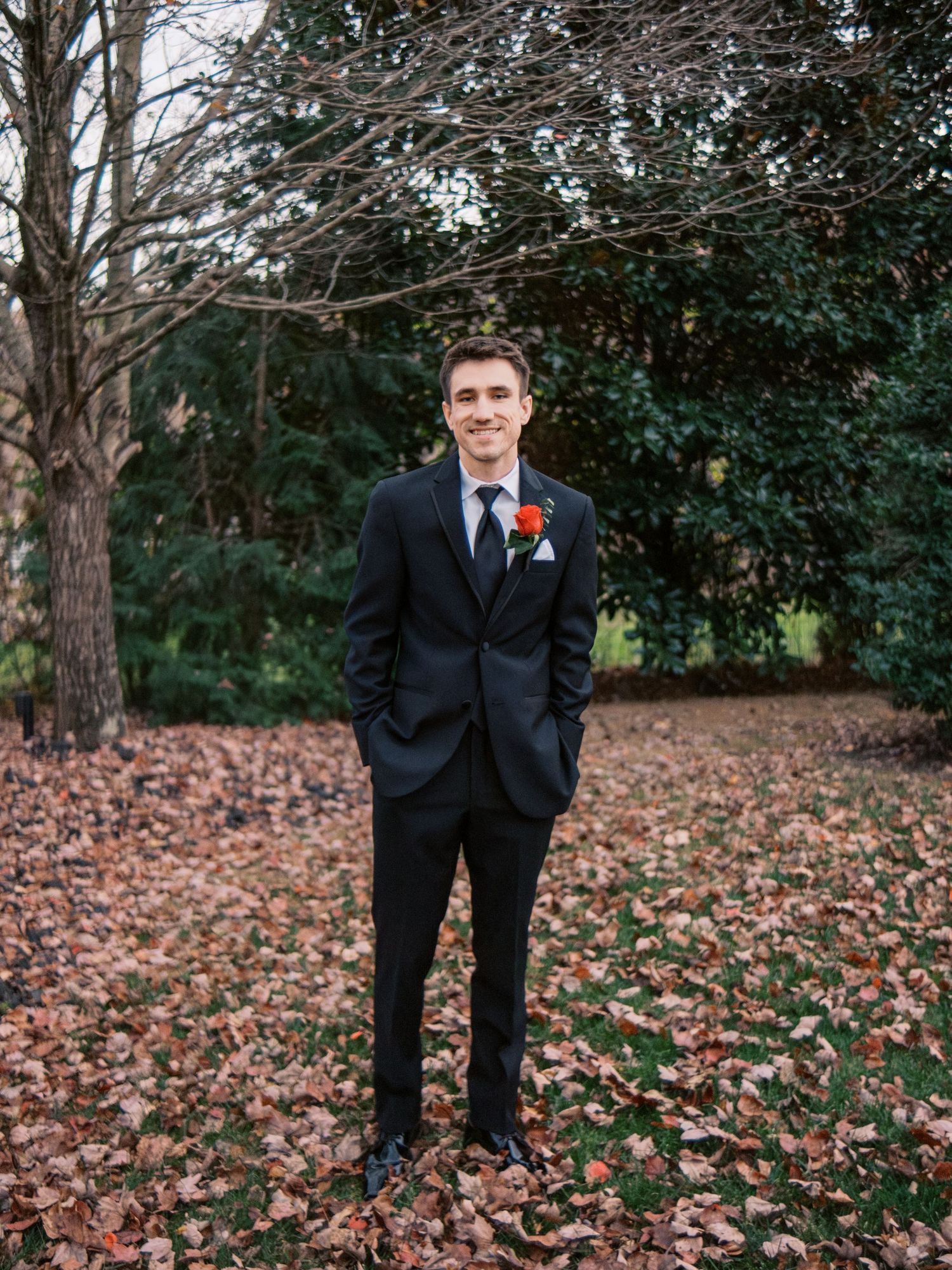 A groom in a black suit and burgundy boutonniere stands on fallen leaves in an autumn garden.