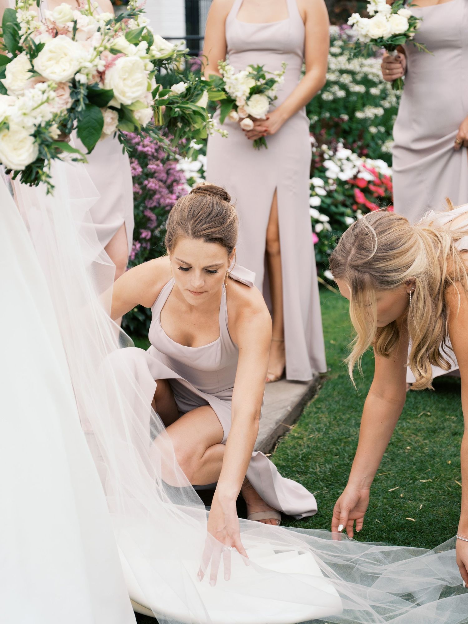 Bridesmaids in blush dresses help arrange the bride's wedding gown on green grass.
