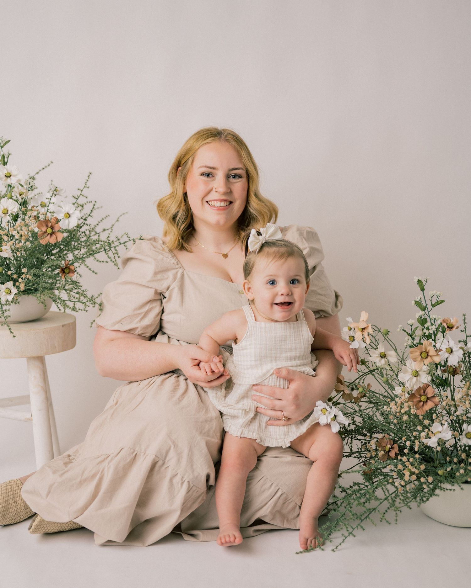 Professional studio portrait with neutral backdrop featuring beige linen dress and white romper amid floral arrangements.