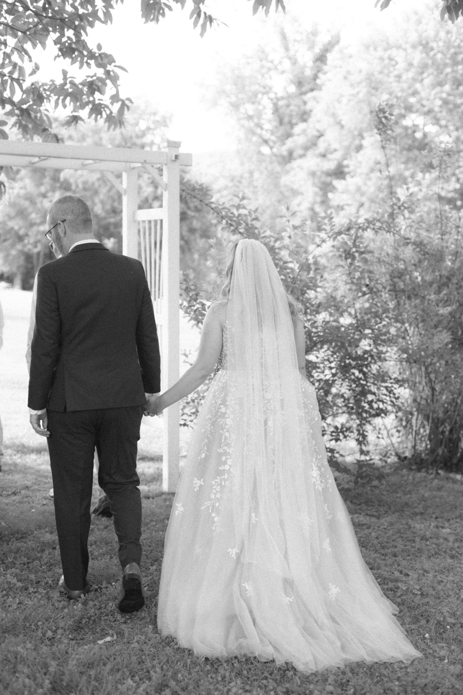 A couple walking through a garden path in formal wedding attire with flowing veil.