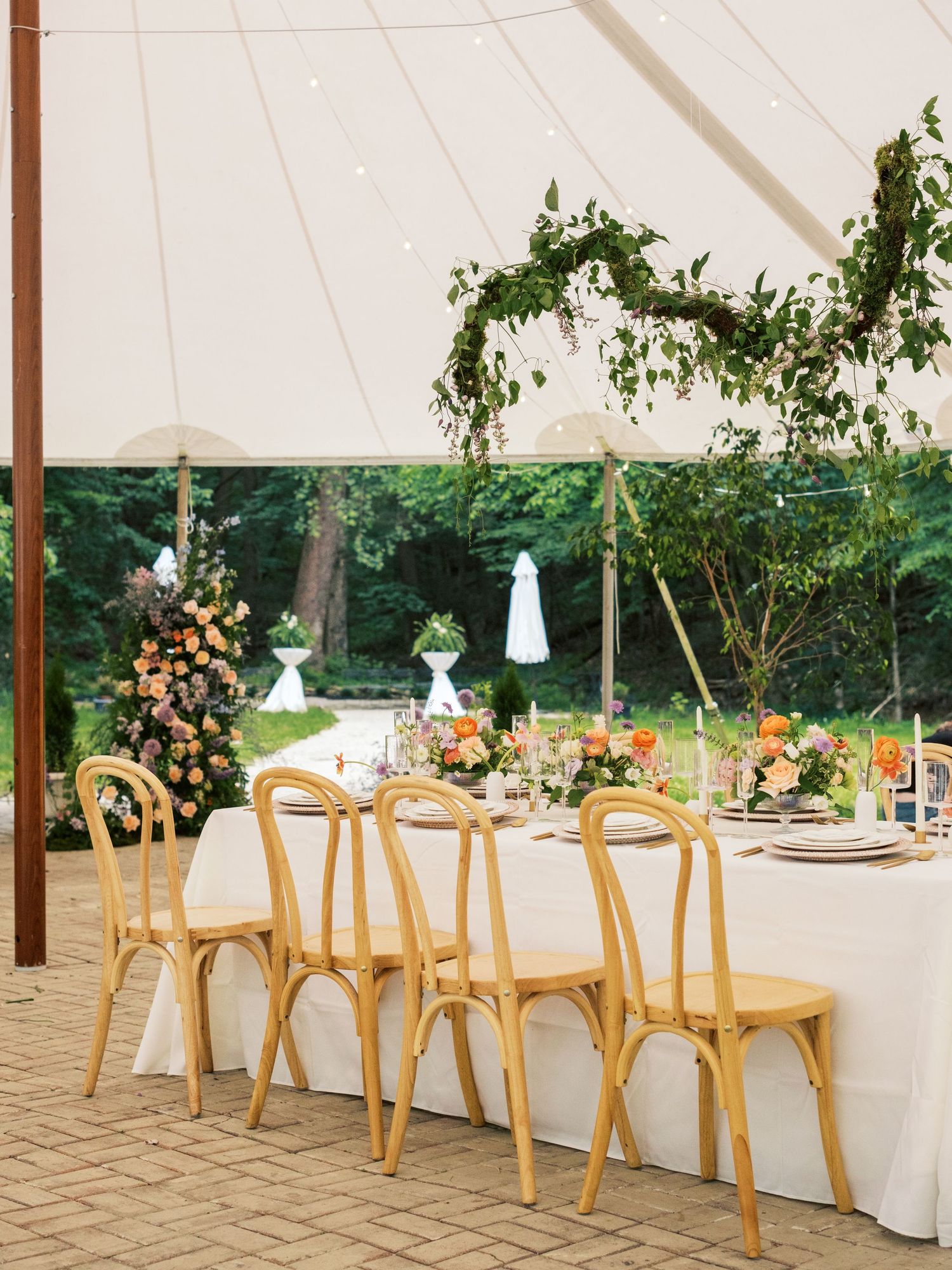 Elegant outdoor reception setup with natural wood chairs and floral arrangements under a white tent canopy.